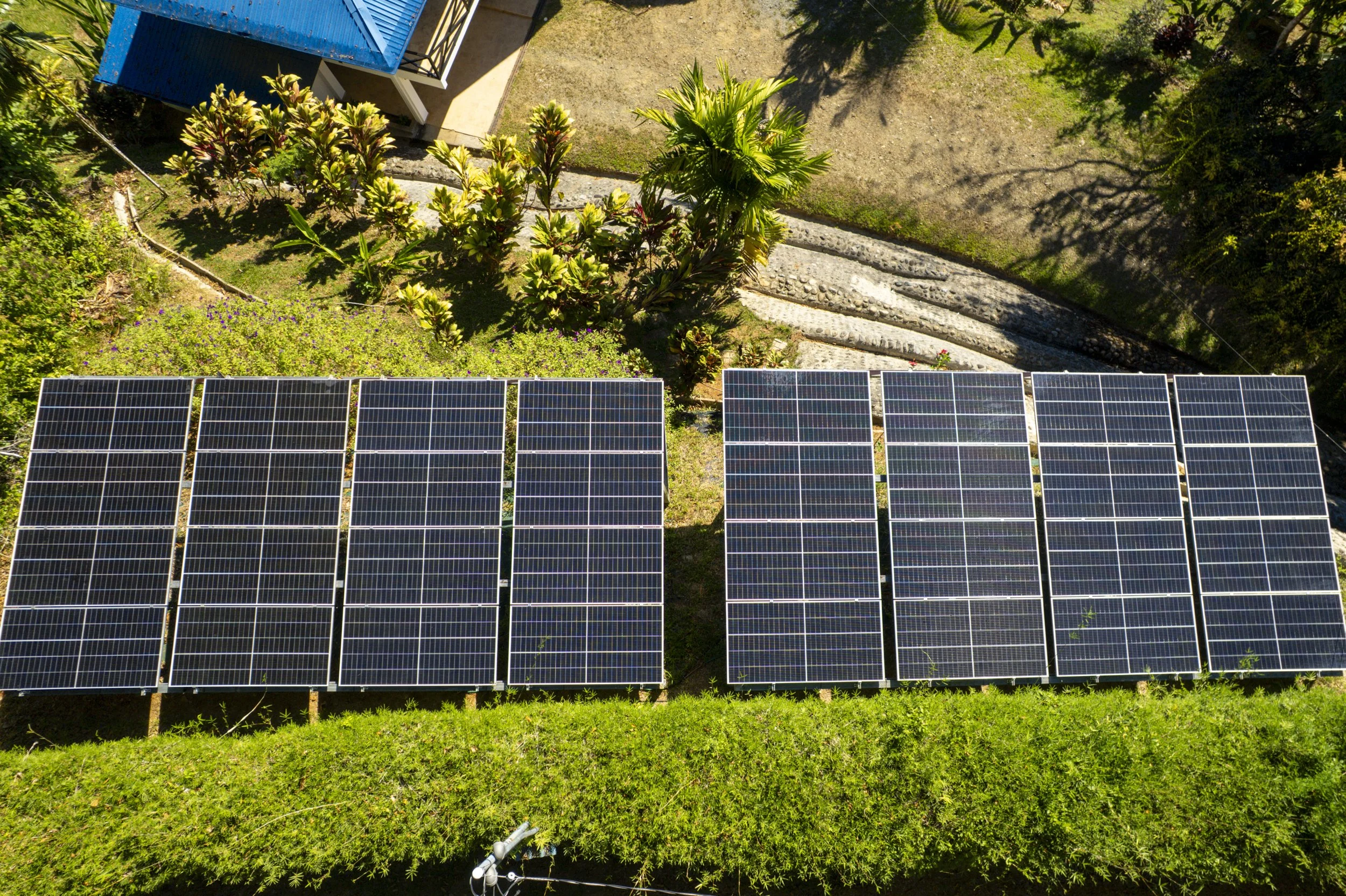 An aerial view of solar panels installed outdoors on grass, with trees and a building nearby.