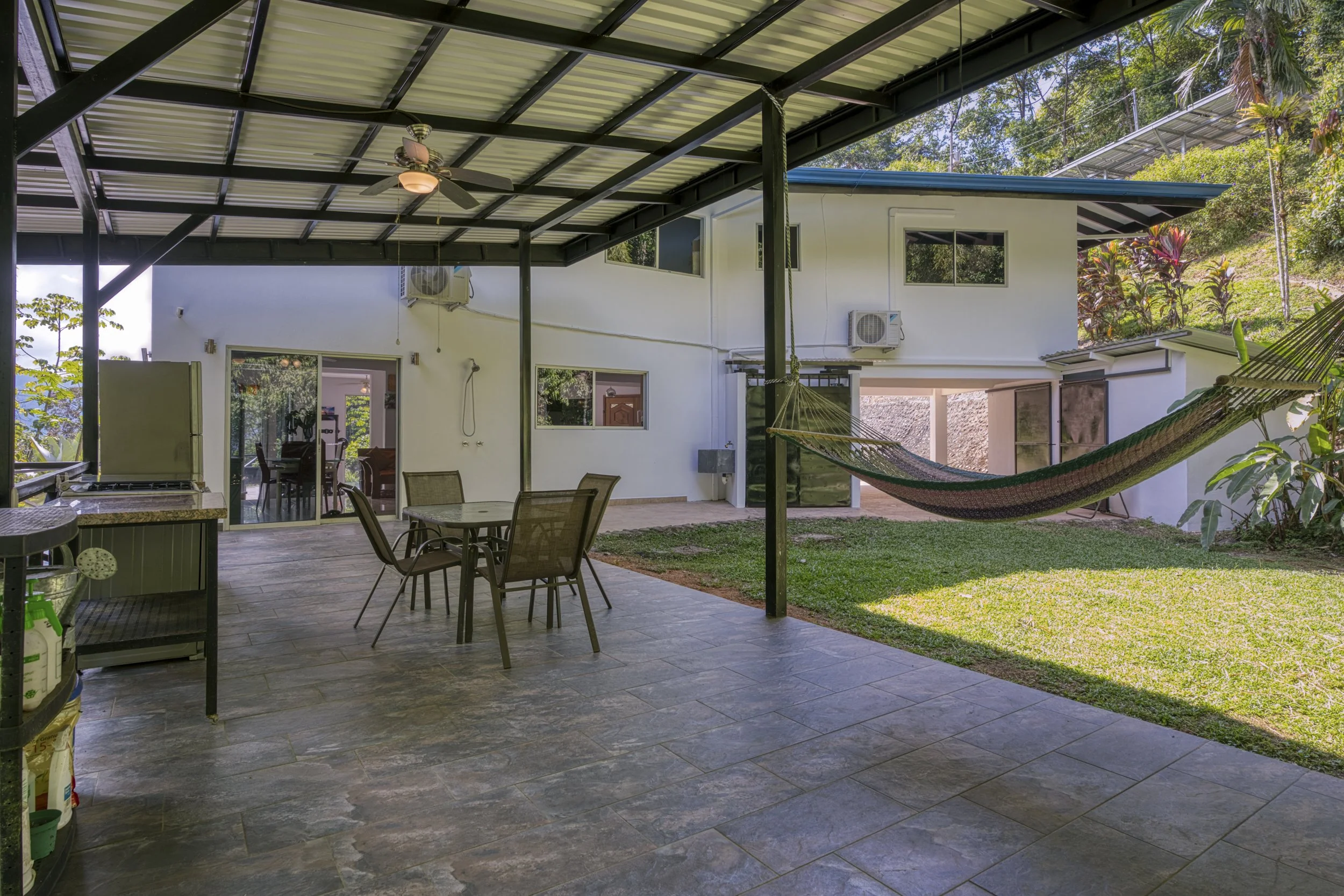 Outdoor covered patio with a ceiling fan, patio furniture including a round table and chairs, a hammock attached to the house, a grassy yard, and a house painted white with air conditioning units and large windows.