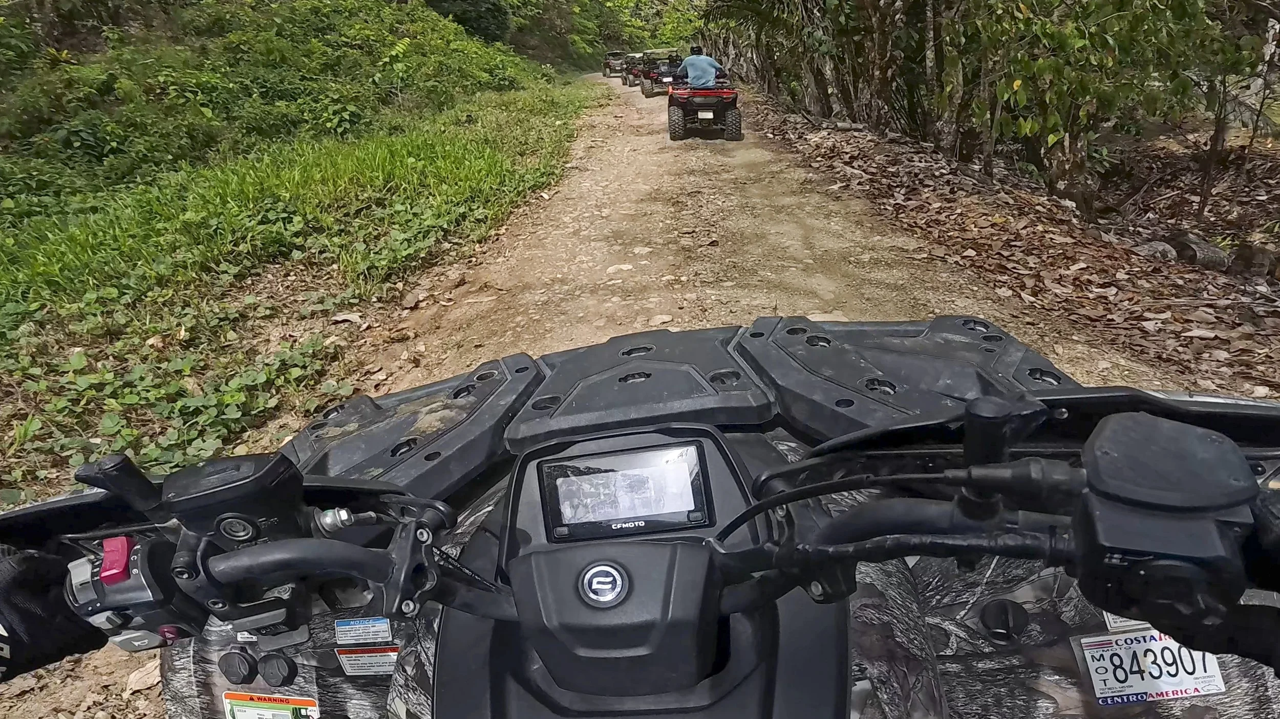 View from a rider on an all-terrain vehicle (ATV) on a dirt trail with multiple other ATV riders following ahead, surrounded by dense green forest.