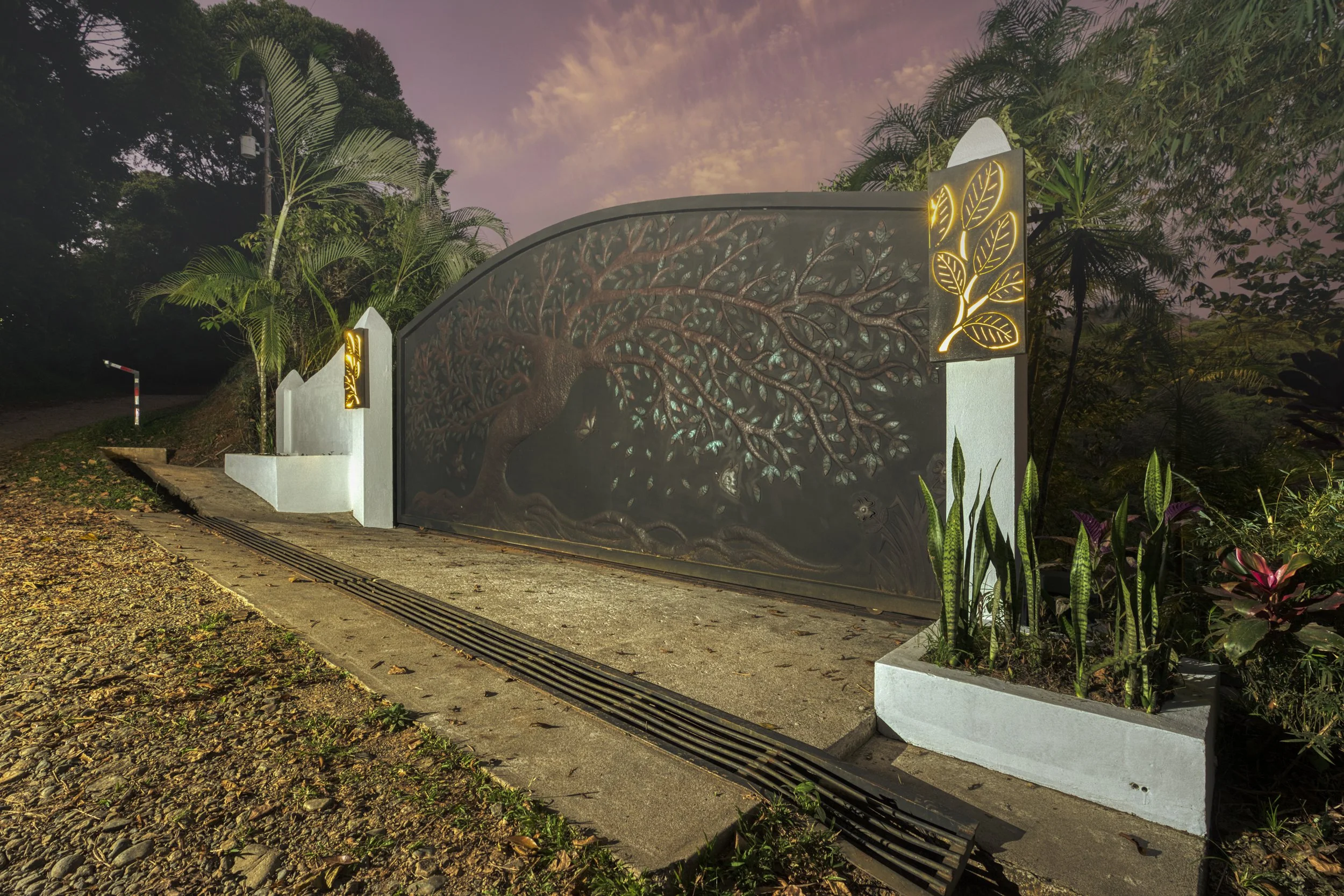 Twilight at the estate entrance reveals a beautiful handcrafted steel gate with the Tree of Life artwork and back-lit floral decorative features on the sides.