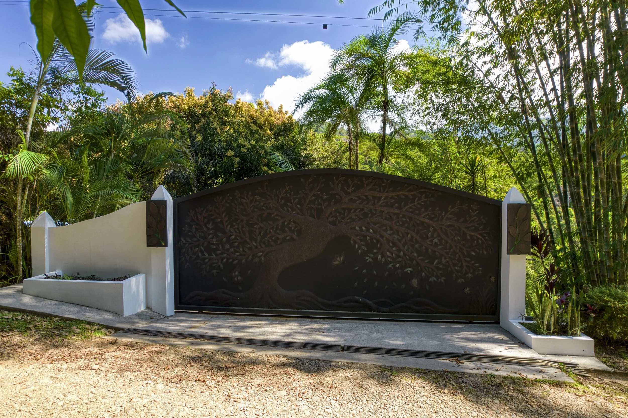 A decorative black metal gate with an intricate tree design, flanked by white concrete pillars with plant motifs, set against lush green tropical foliage and a partly cloudy blue sky.