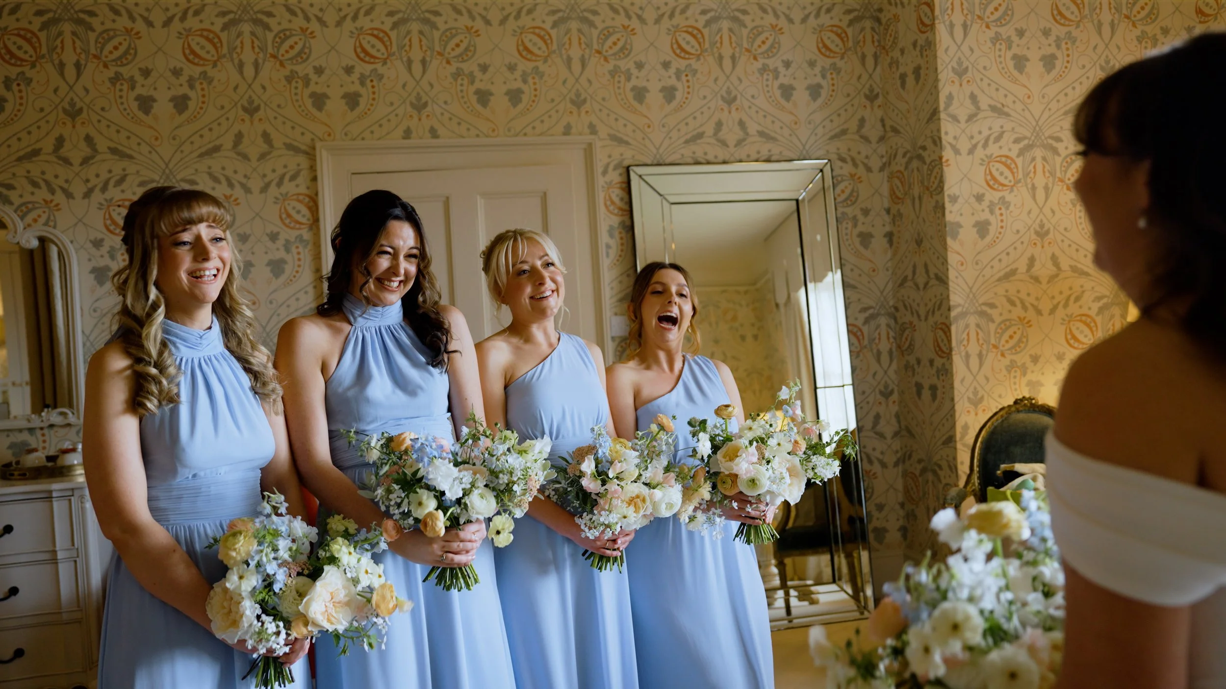 Four bridesmaids in matching light blue dresses holding bouquets in a room with patterned wallpaper, as they smile at a bride in an off-the-shoulder white dress holding a bouquet.