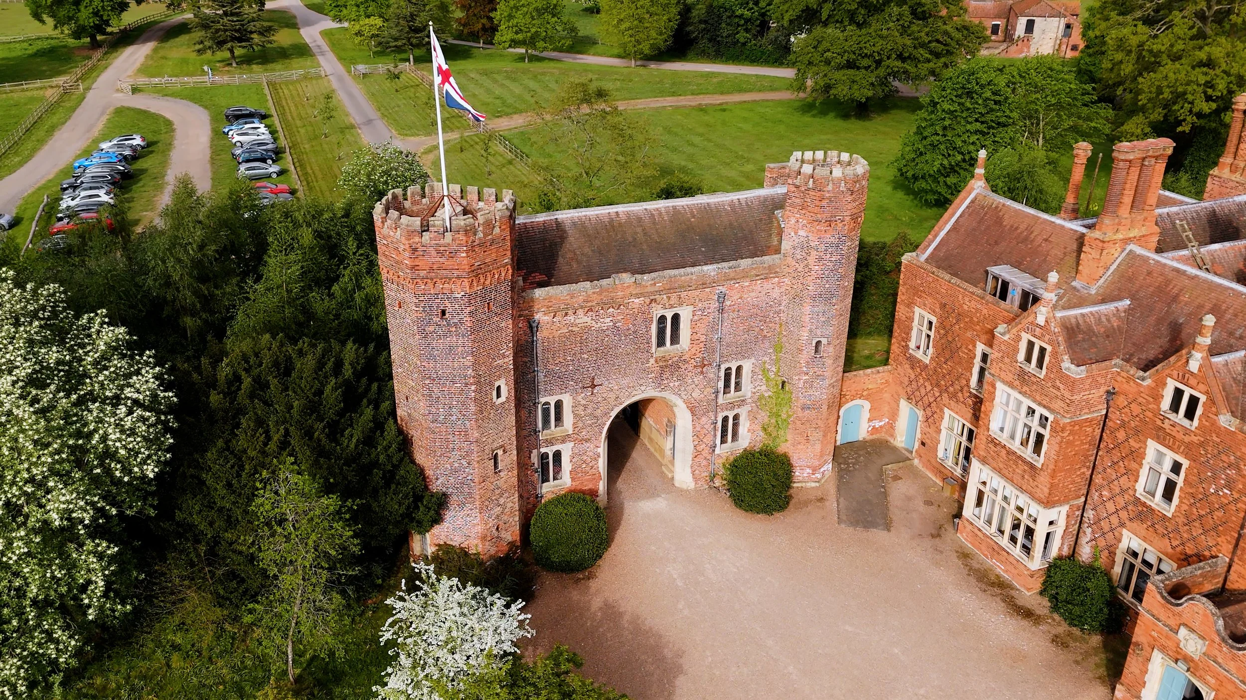 Aerial view of a historic brick castle with a clock tower and a flag on top, surrounded by greenery, trees, a gravel pathway, and a parking lot with cars.
