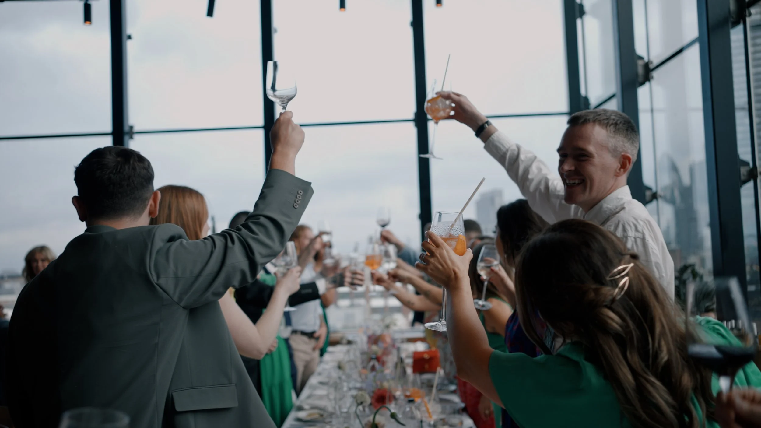 People raising glasses during a celebration or toast at a party in a modern indoor venue with large windows and city skyline view.