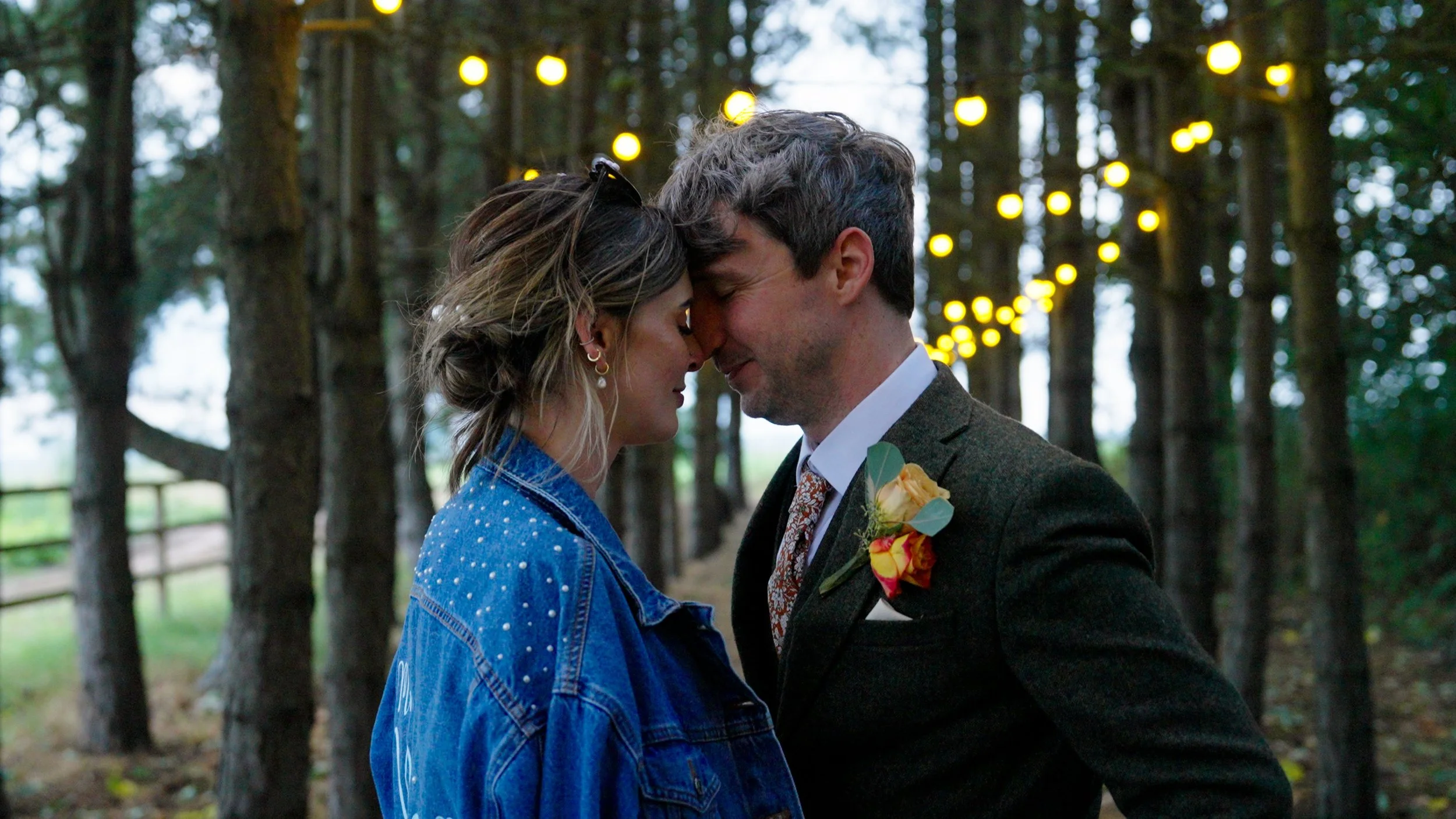 A couple standing close, their foreheads touching, in a wooded area with string lights hanging above. The man is dressed in a suit with a boutonniere, and the woman is wearing a blue jacket with pearl accents.