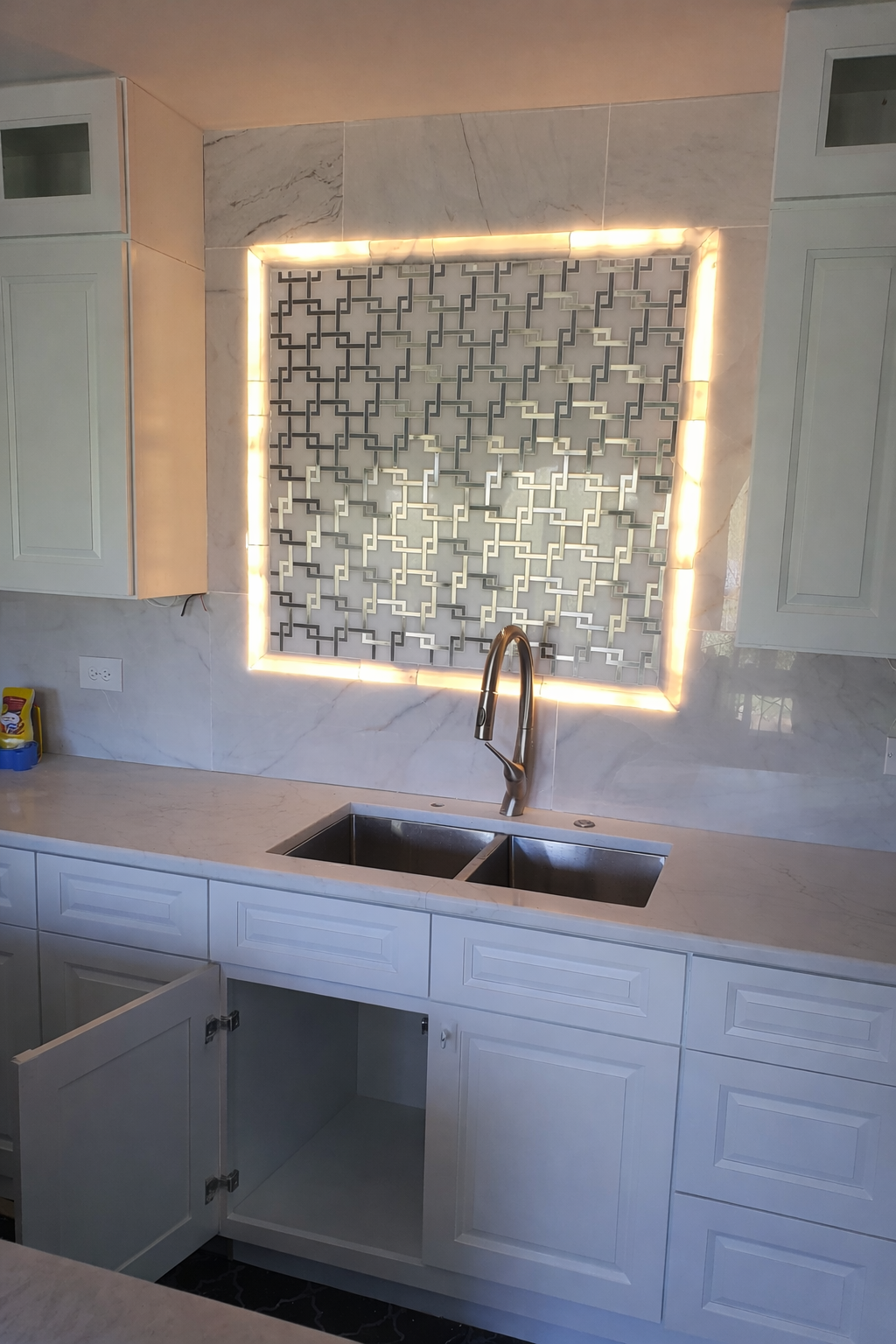 Modern kitchen with white cabinets, marble countertop, stainless steel double sink, and a decorative tile backsplash with backlighting.