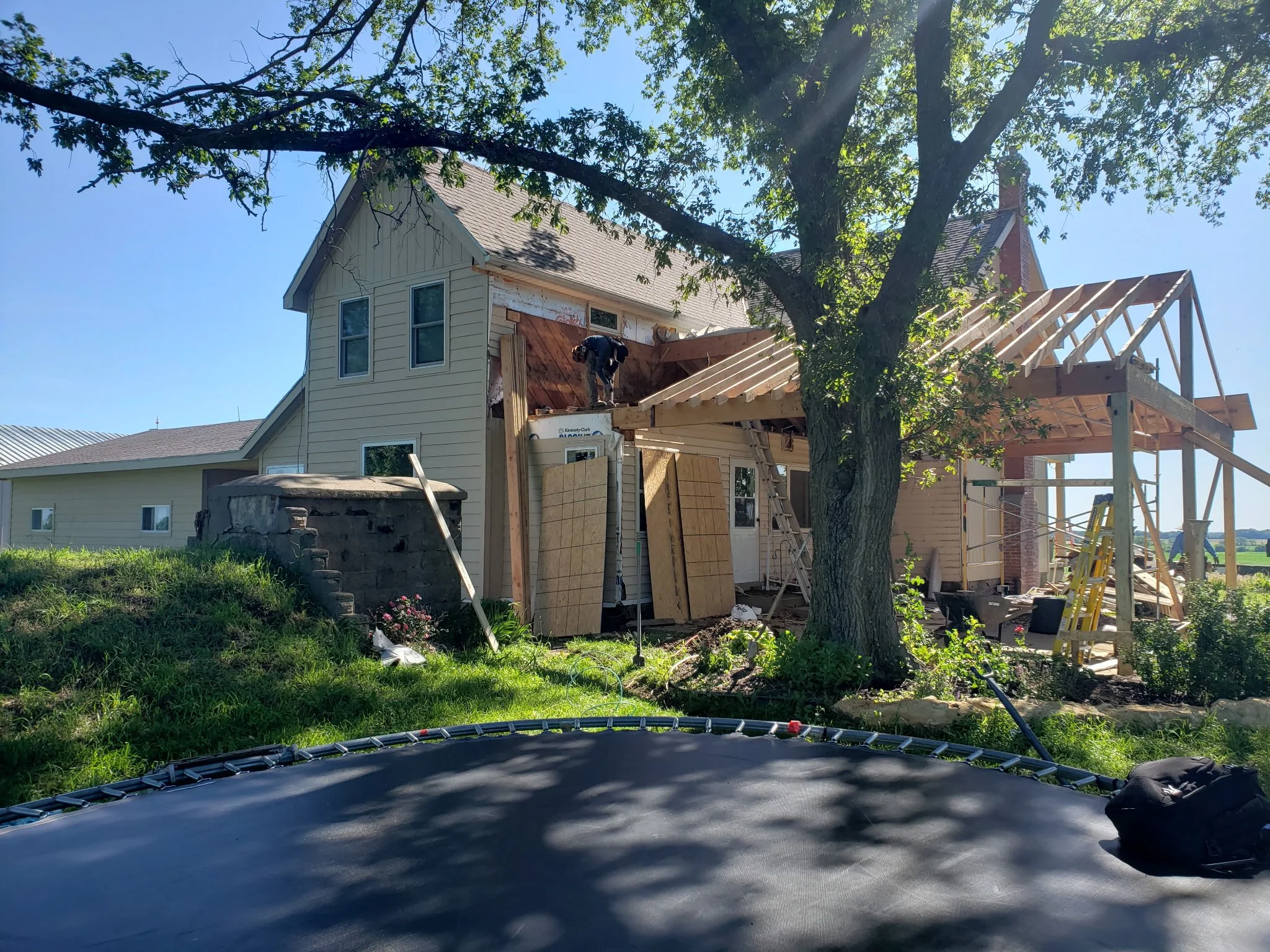 House under construction with a worker on the roof, wooden framing for a new extension, a large tree in the yard, and a trampoline in the foreground.