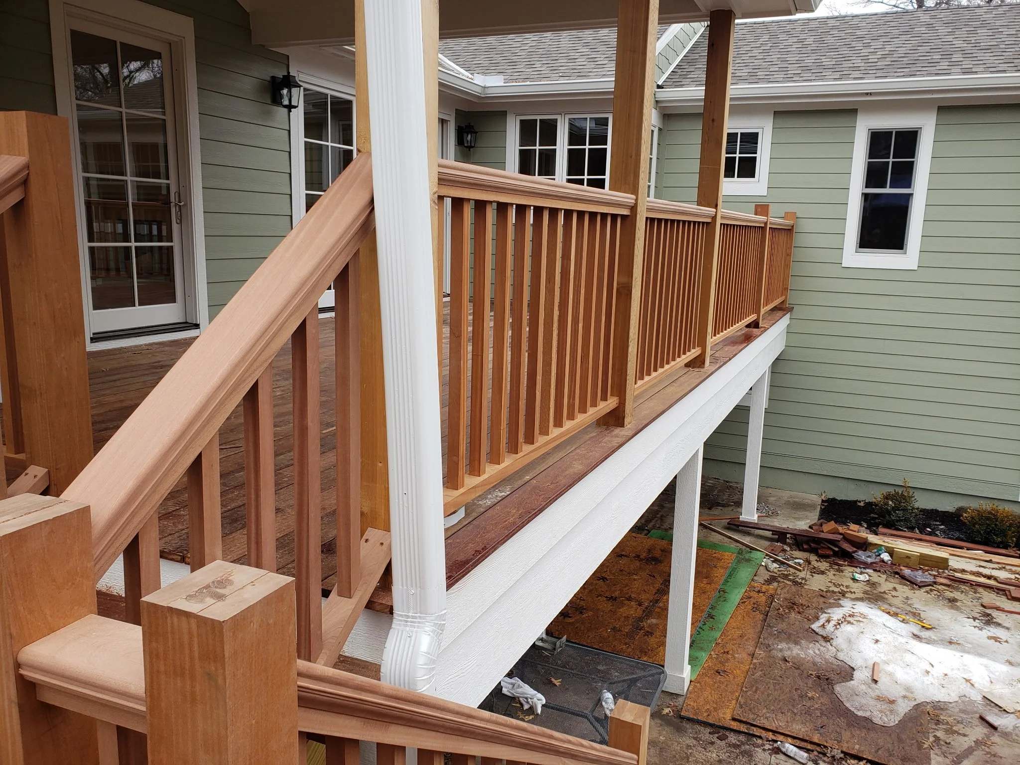 A newly built wooden deck with railing attached to a house, with construction materials and tools visible below, and snow on the ground.