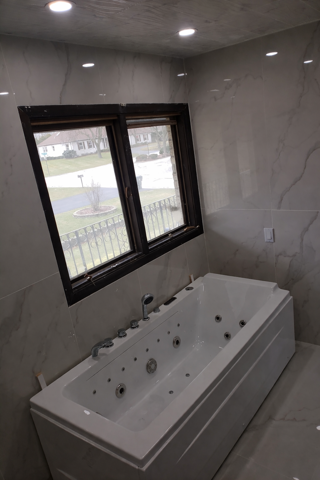 A bathroom with a whirlpool bathtub under a window, marble walls, and built-in lighting on the ceiling.