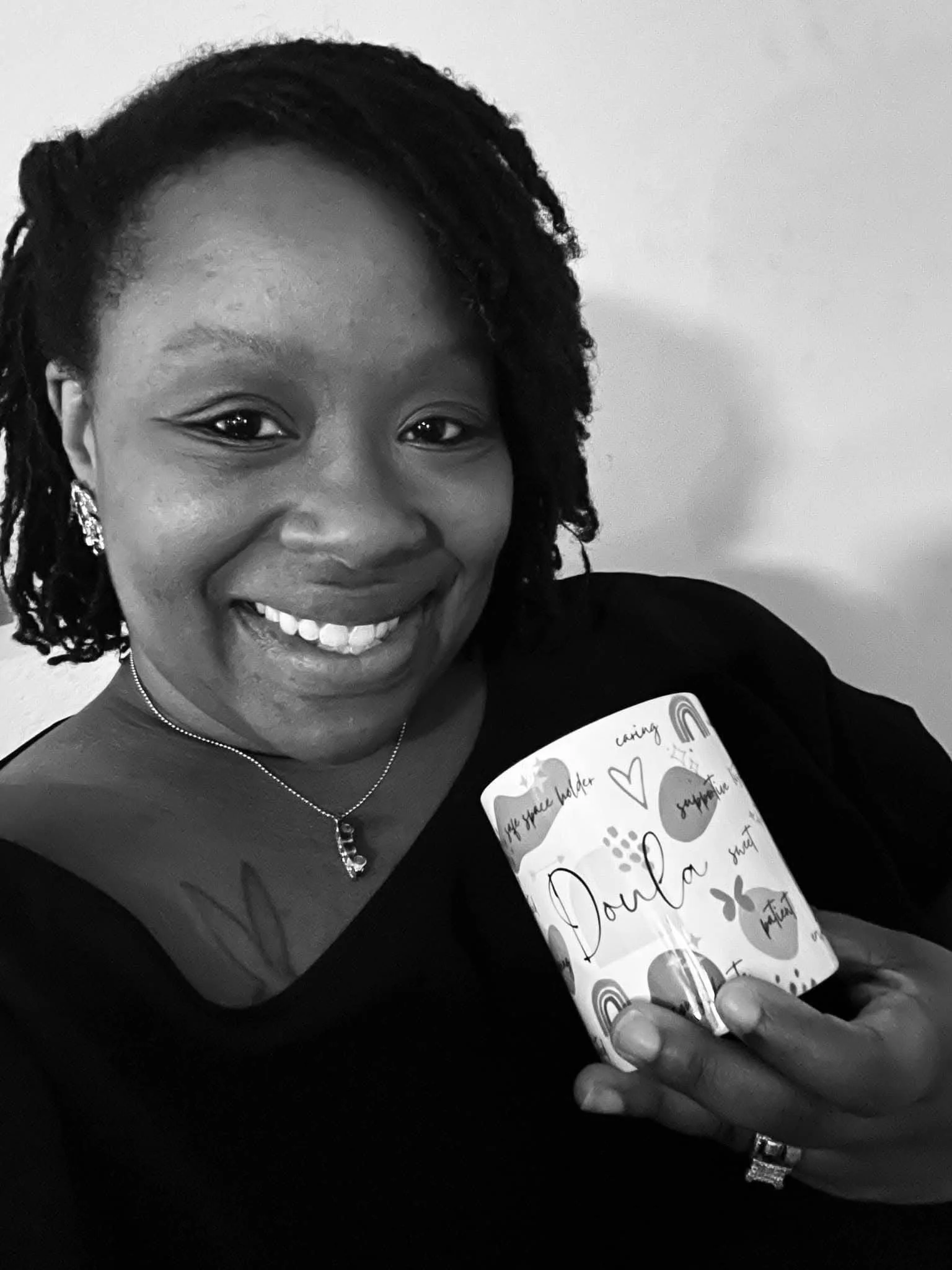 CBC community doula, Shieila, a black woman, smiling wearing a black shirt and holding a mug that says “doula”.