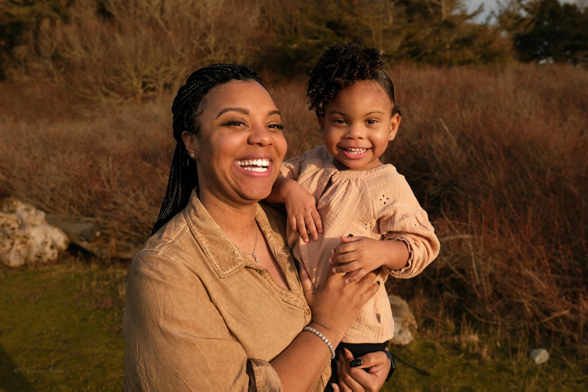 A woman and a young girl smiling outdoors, with the woman holding the girl in her arms. The background shows a natural setting with trees and bushes.