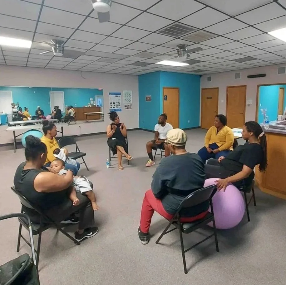 Group of black people and one breastfeeding baby sitting in chairs in a circle during a breastfeeding support class.