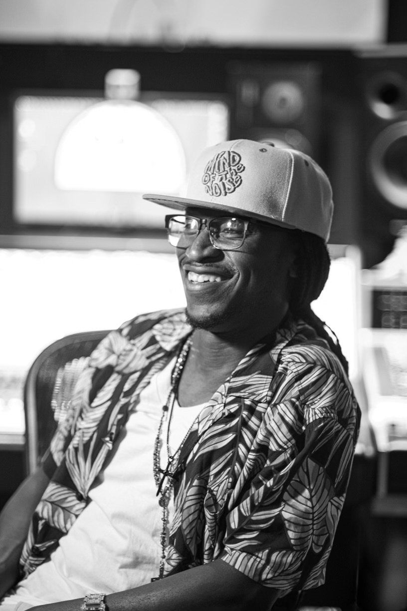 A smiling man wearing glasses, a patterned shirt, a baseball cap with a logo, and jewelry, sitting in a music studio surrounded by audio equipment.