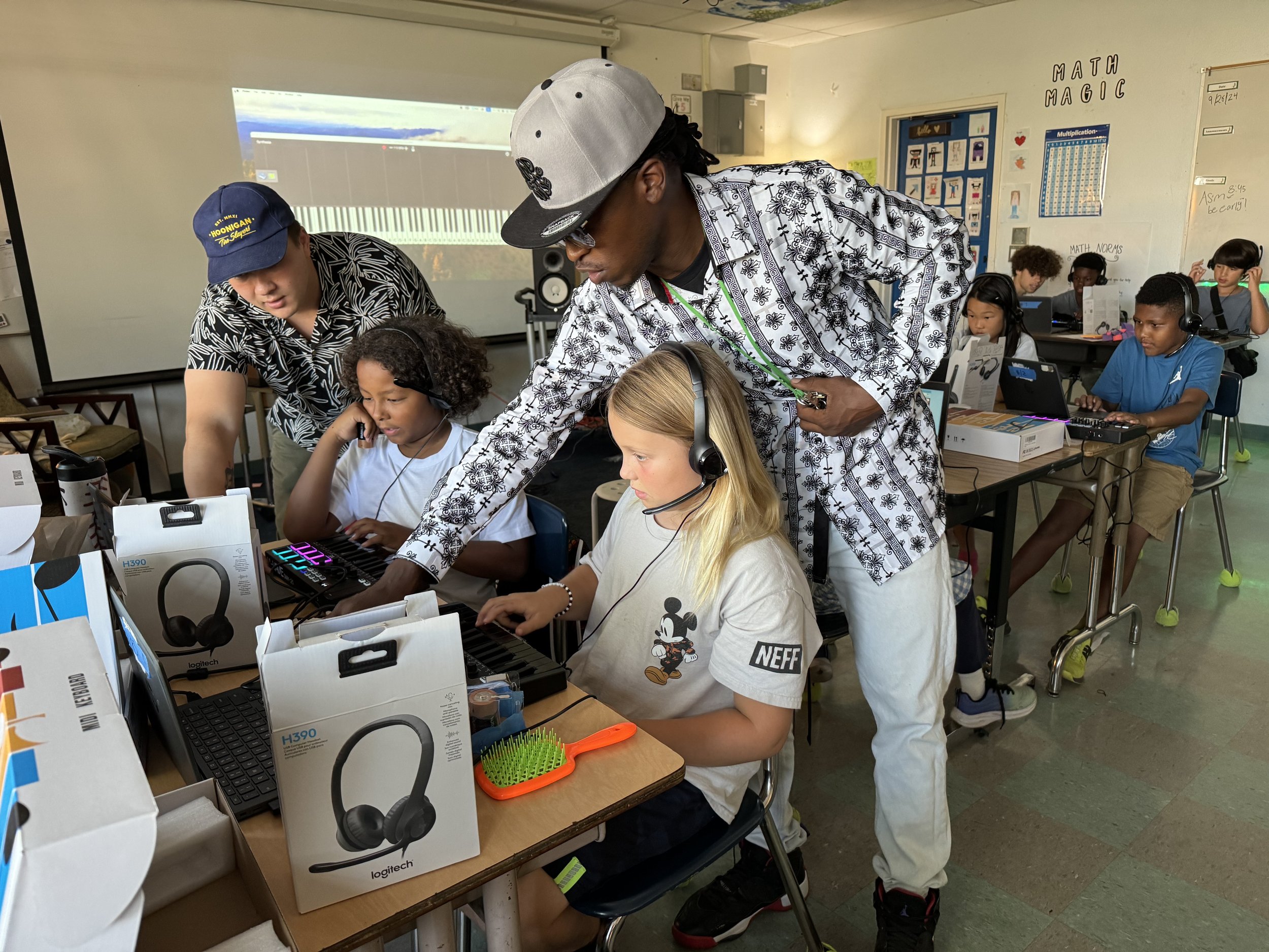 Kids in a classroom working on laptops and electronic music equipment, with teachers assisting, surrounded by educational posters and a whiteboard.