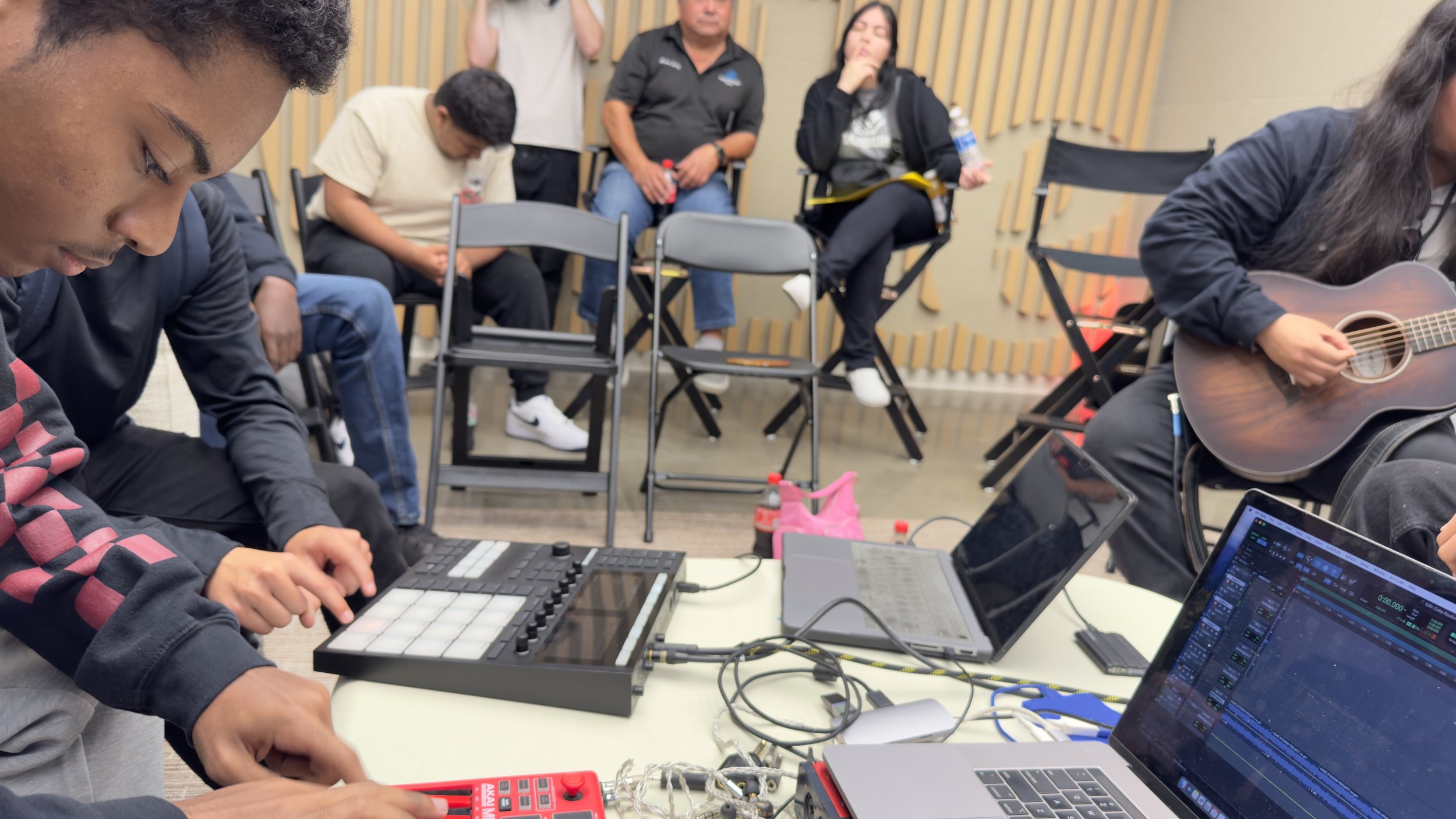 People in a music studio with laptops, audio equipment, and a person playing guitar.