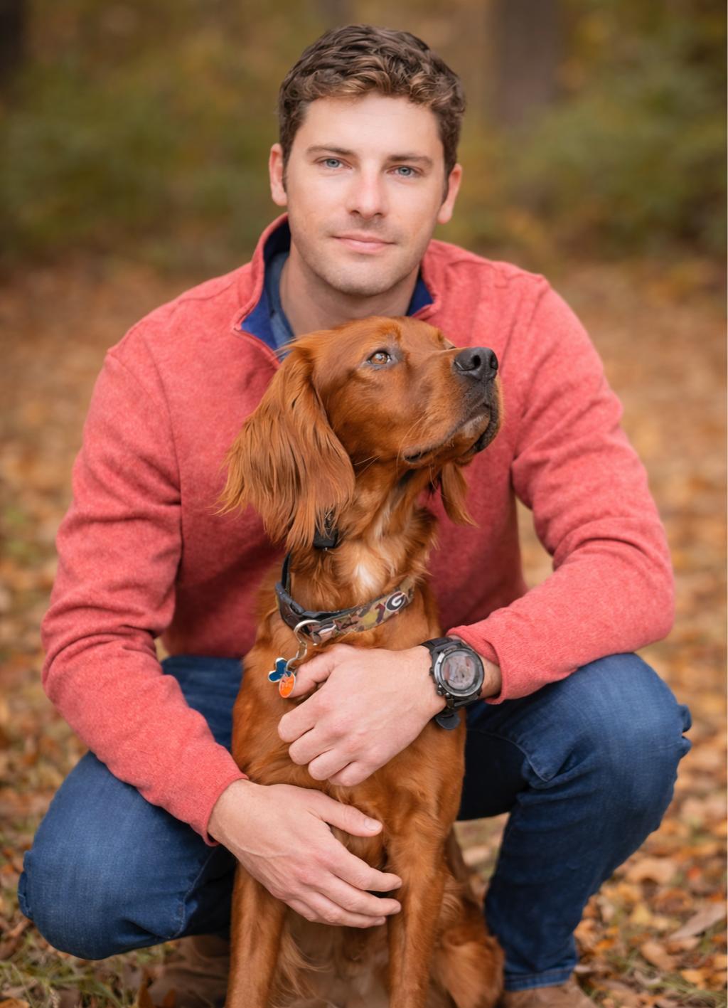 A man in a red jacket squats outdoors, holding a brown dog with floppy ears, in an autumnal setting.