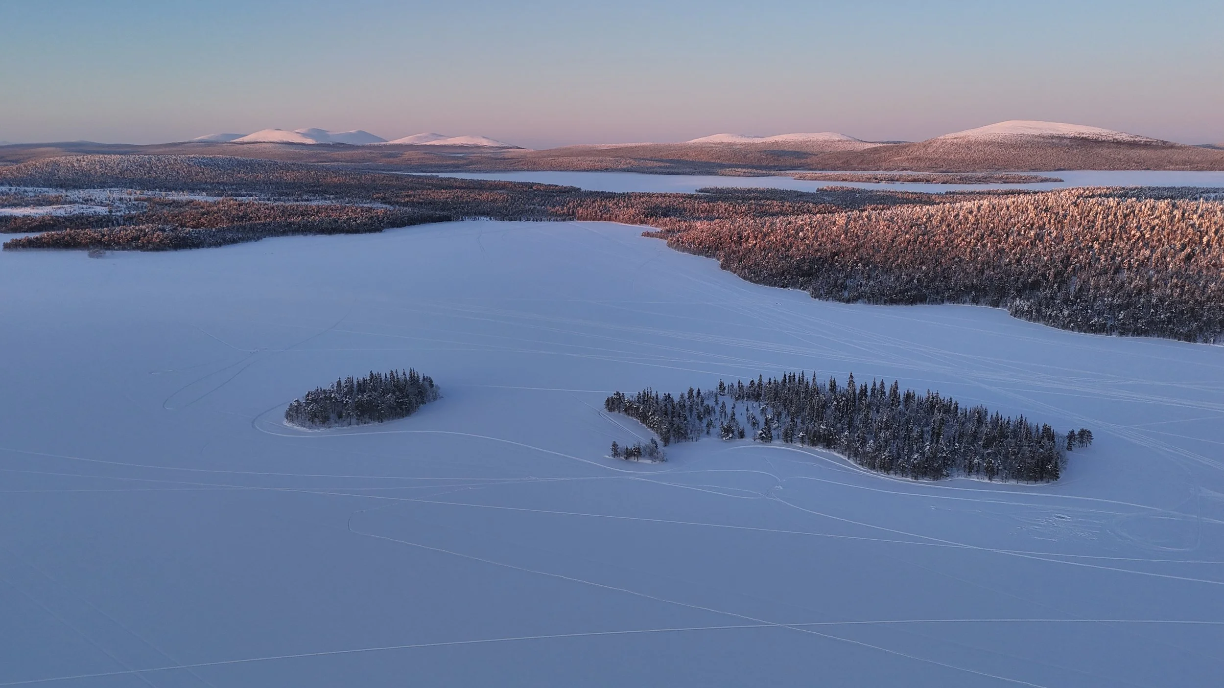 Snow-covered frozen lake in Lapland with two small pine tree islands, surrounded by dense forest and distant snow-capped mountains under a clear sky. Aerial view shot by drone.