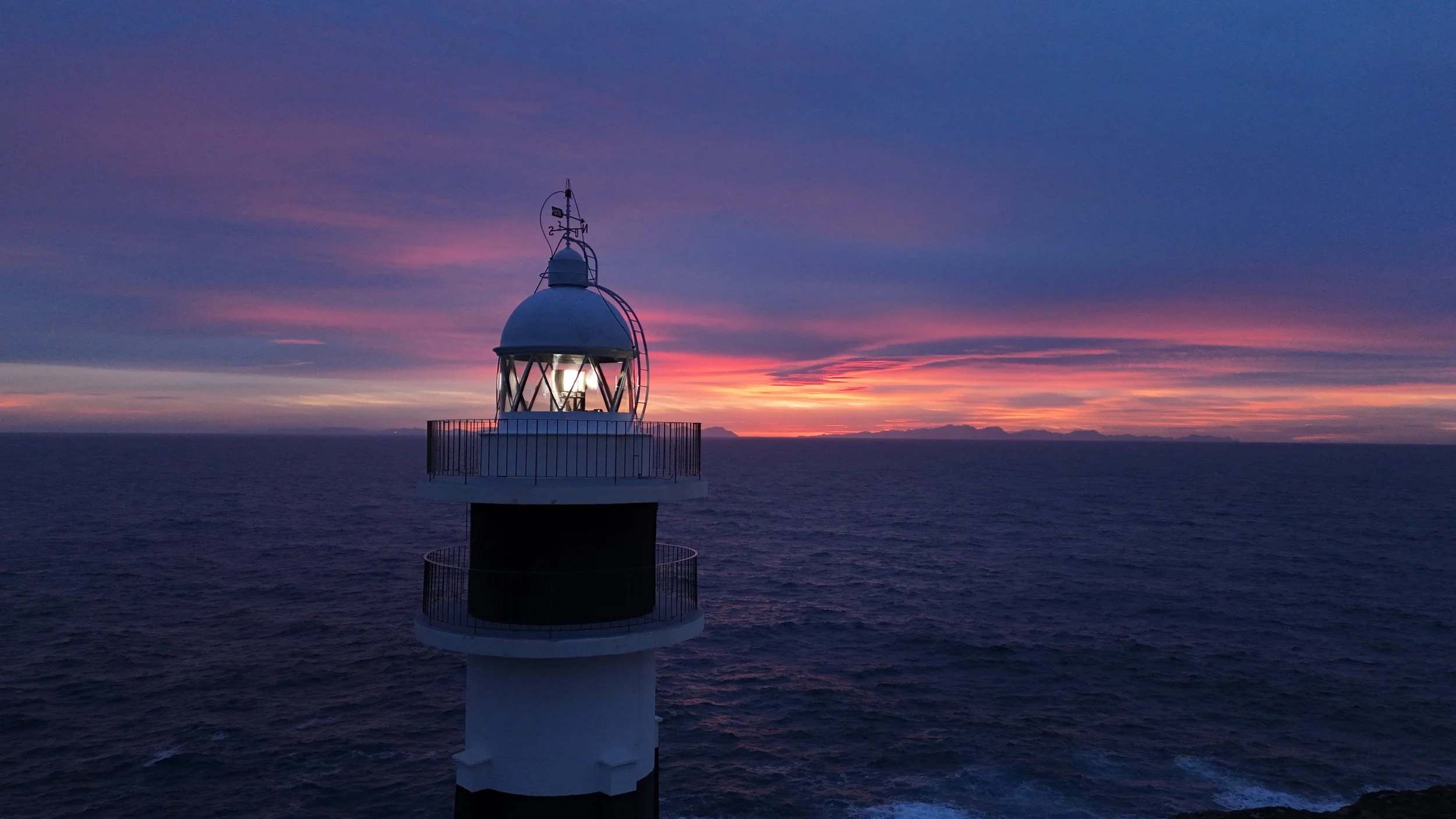 A lighthouse stands on the edge of the ocean during sunset, with a colorful sky of pink, purple, and orange hues. Aerial view shot by drone. Cap d'Artrutx en Menorca.