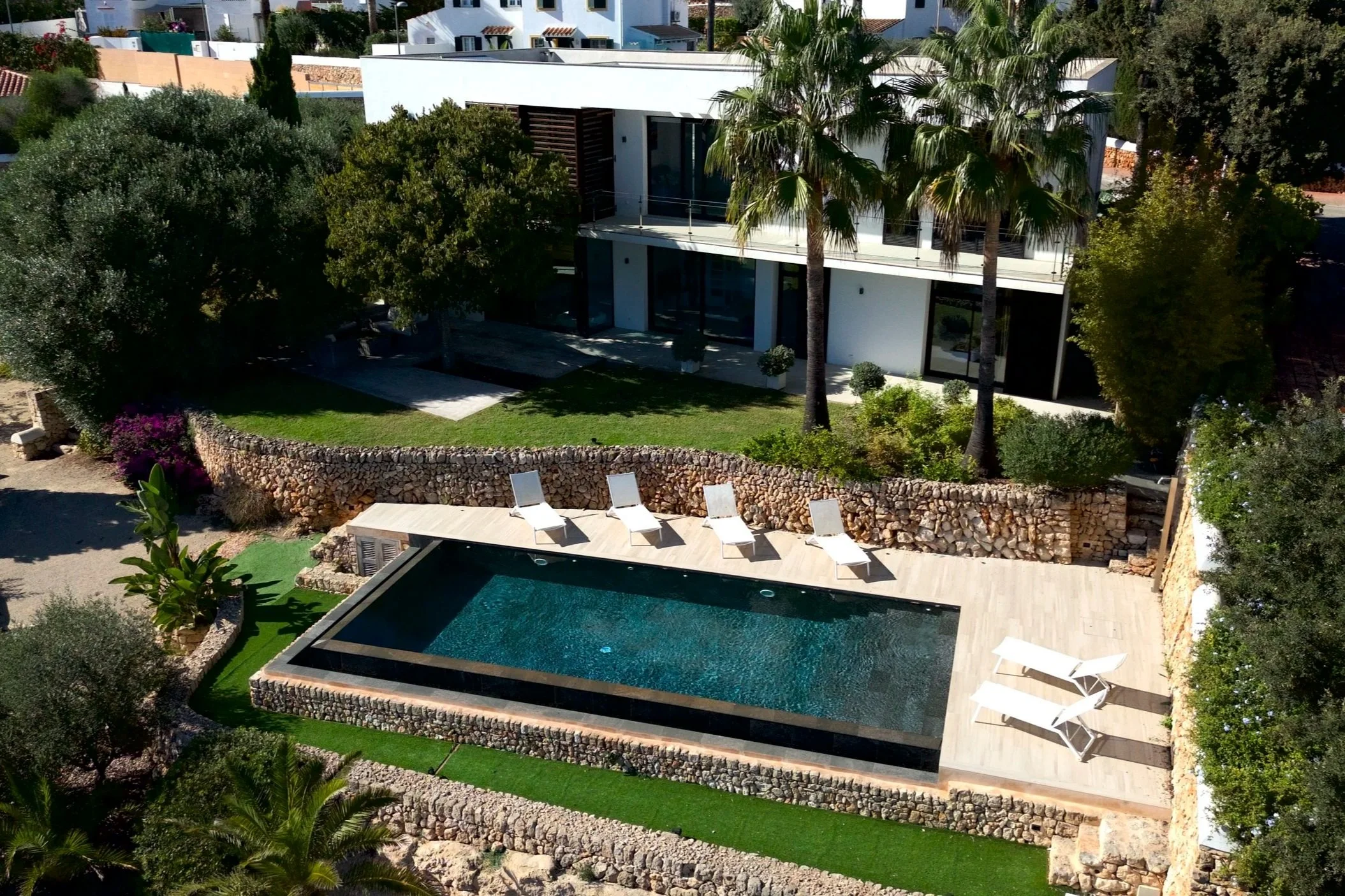 Drone aerial photography of a modern two-story house in Menorca with large glass windows and a balcony, surrounded by palm trees and lush greenery, featuring a rectangular swimming pool with four white lounge chairs on a wooden deck.