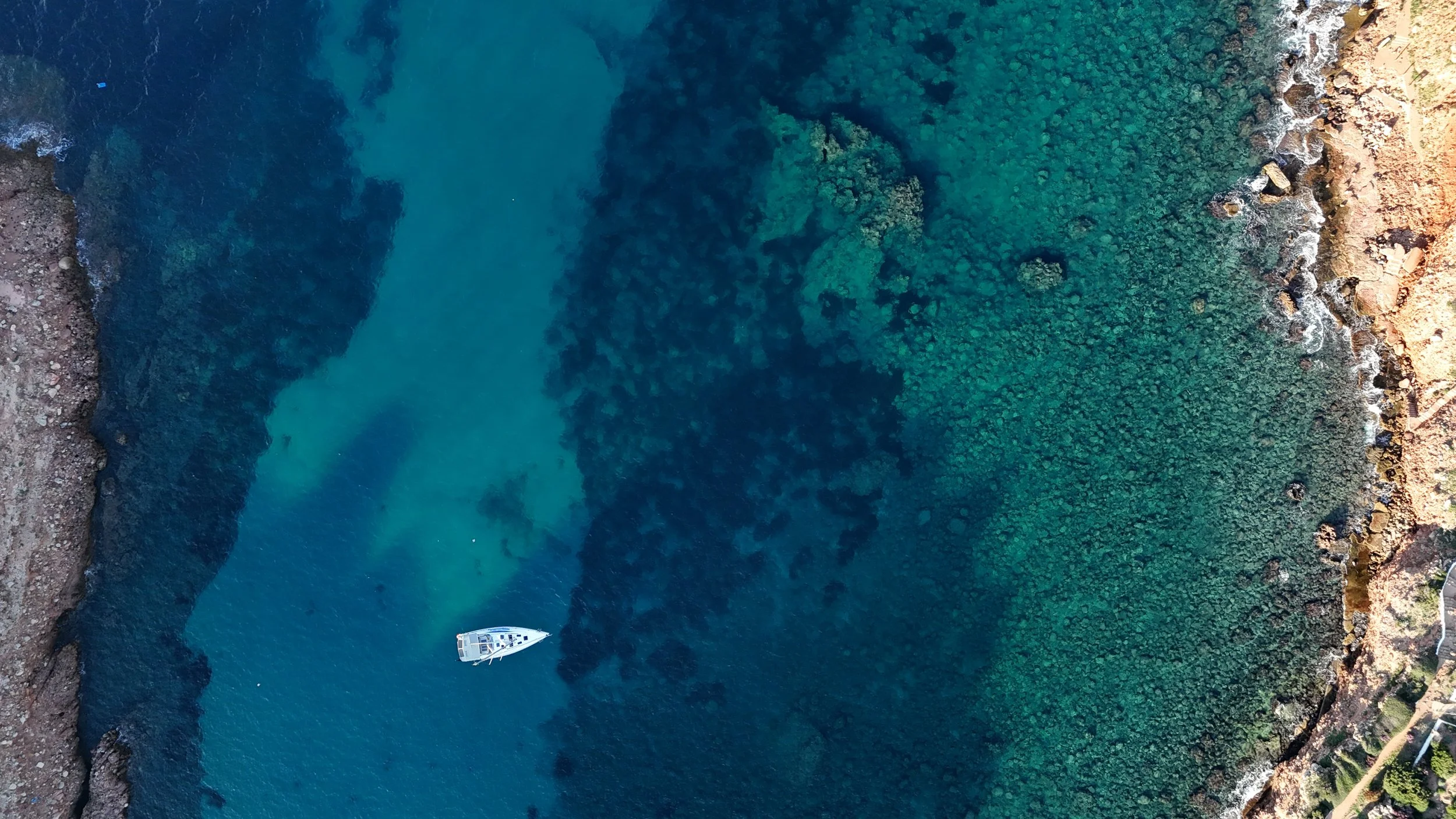 An aerial view shot by drone of a white boat anchored in clear, blue-green waters near a rocky shoreline with lush green vegetation in Menorca.