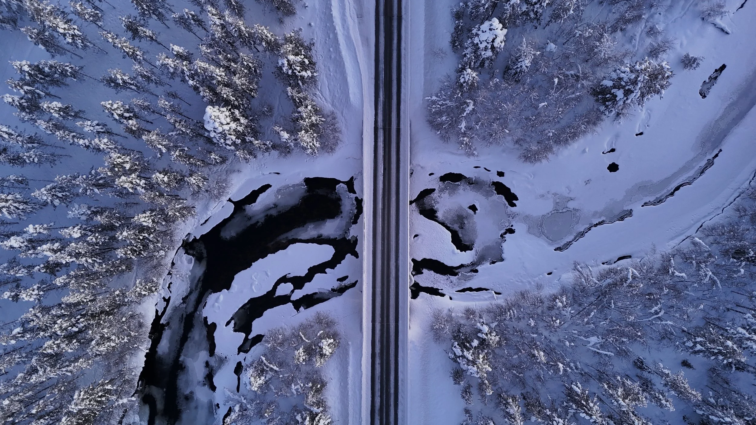 An aerial view shot by drone of a snow-covered forest and a partially frozen river, with a bridge crossing the river.