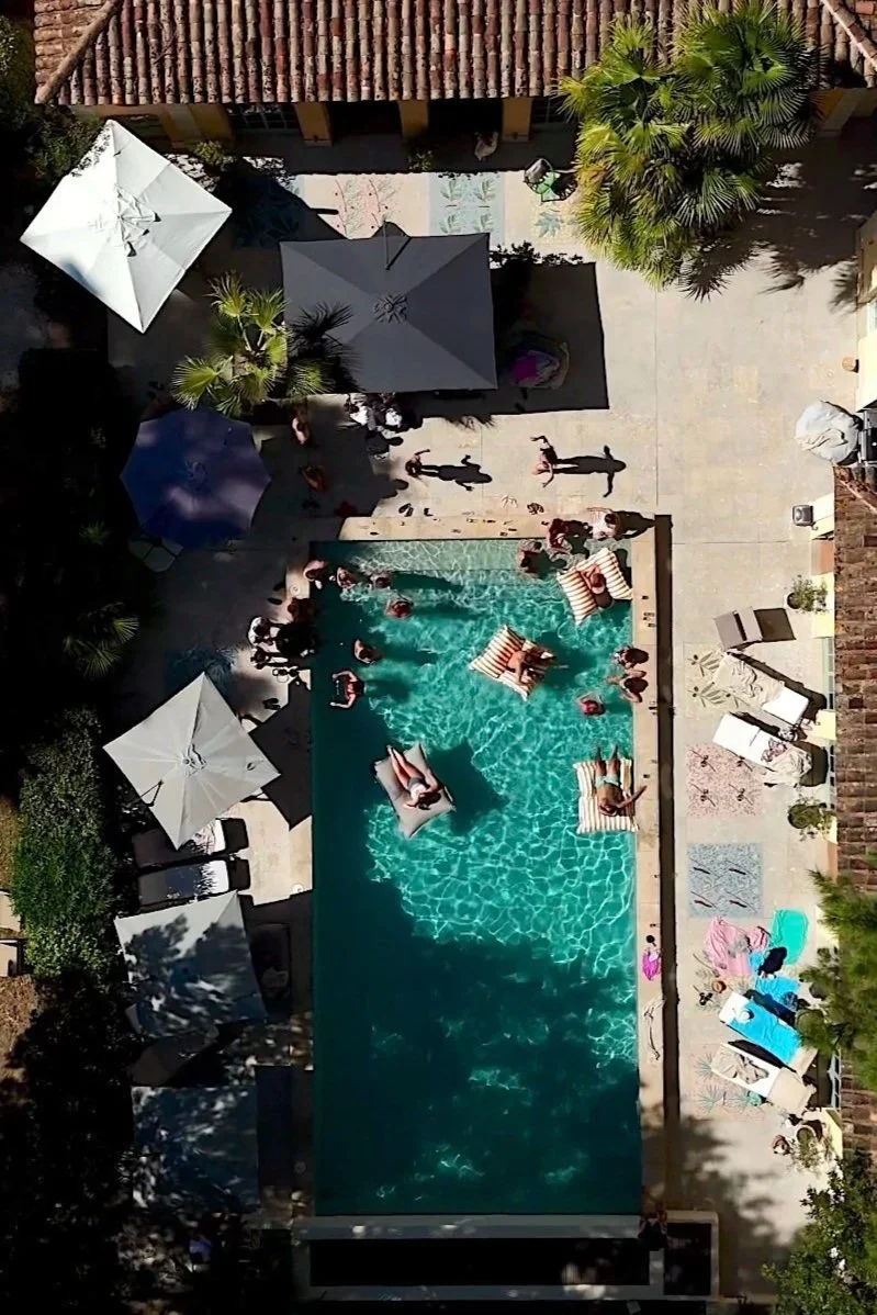 A backyard swimming pool area viewed from above, with people swimming and lounging on pool floats, and shaded by umbrellas and trees. Drone photography taken in Menorca
