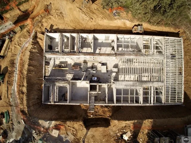 Aerial view by drone of a large house under construction with visible framing, interior walls, and an unfinished roof. Construction equipment and materials are scattered around the site.