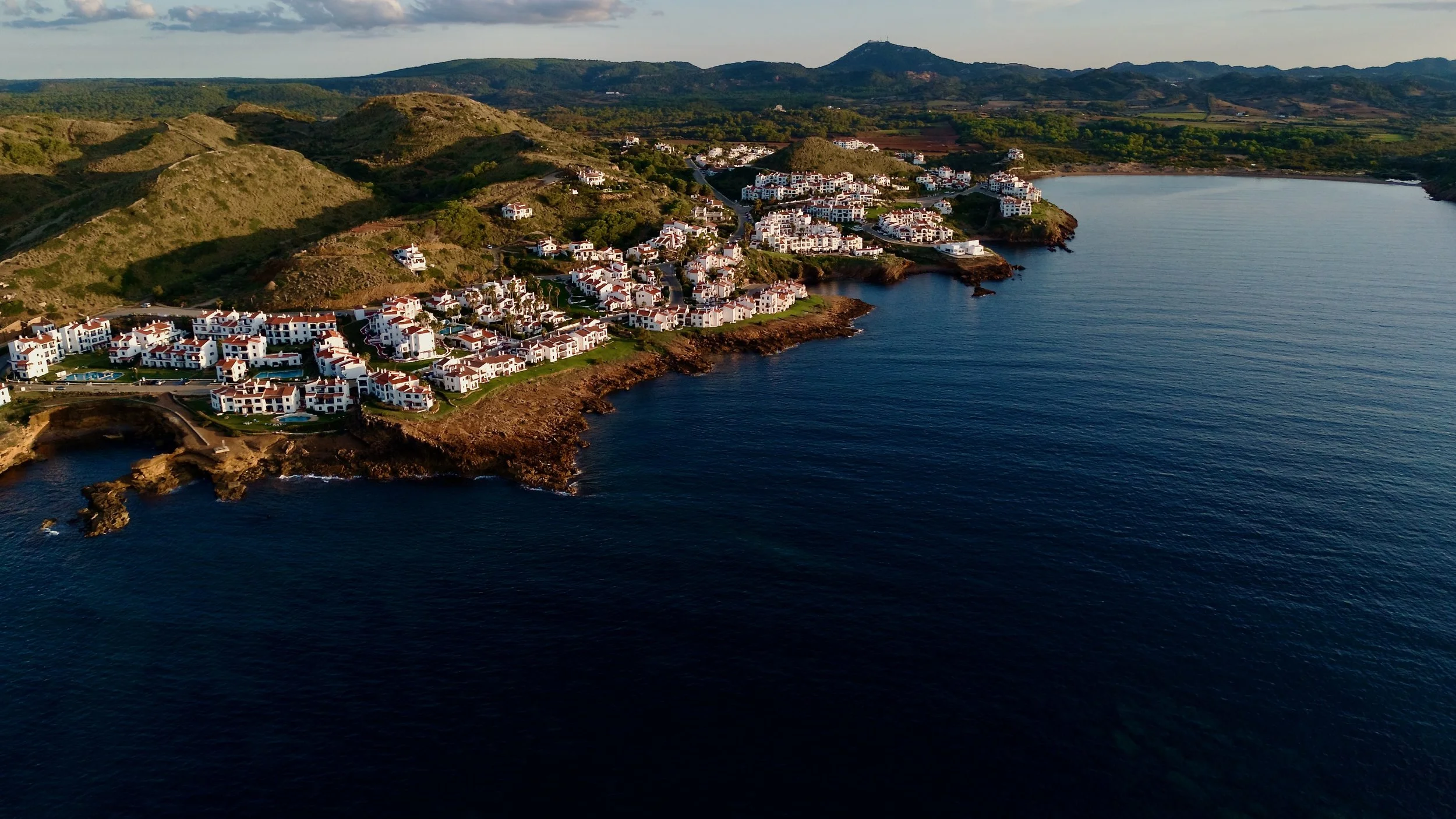 Aerial view shot by drone of a coastal residential area in Menorca with white buildings and red rooftops along rocky shores, surrounded by green hills and deep blue ocean waters.