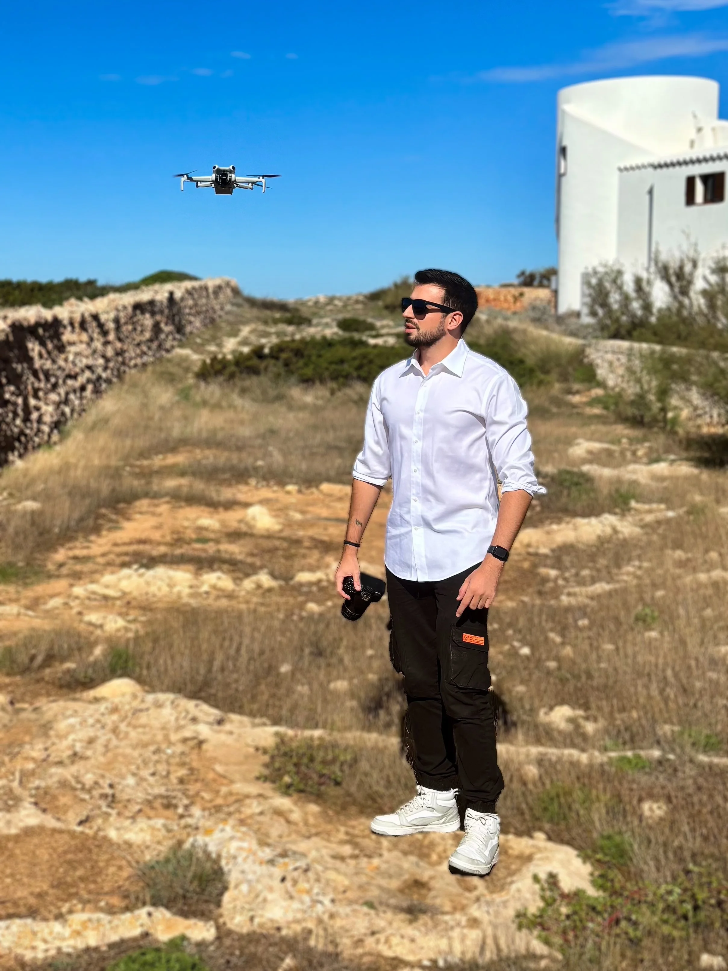 A man in a white shirt and black cargo pants standing outdoors in Menorca, holding a camera, with a drone flying above him against a blue sky, in a rocky and grassy landscape with white buildings in the background.