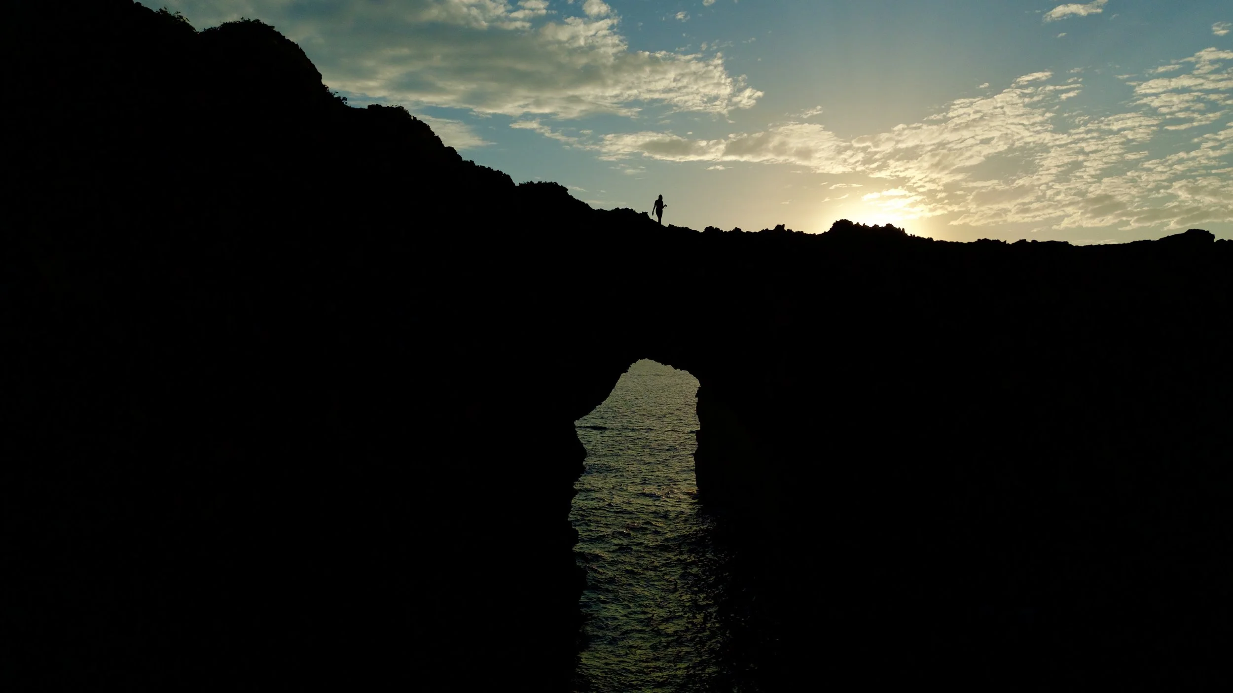 Sunset over a rocky coastal archway in Menorca with a silhouette of a person standing on the cliff above, with calm water reflecting the arch and sky with scattered clouds. Aerial view shot by drone.