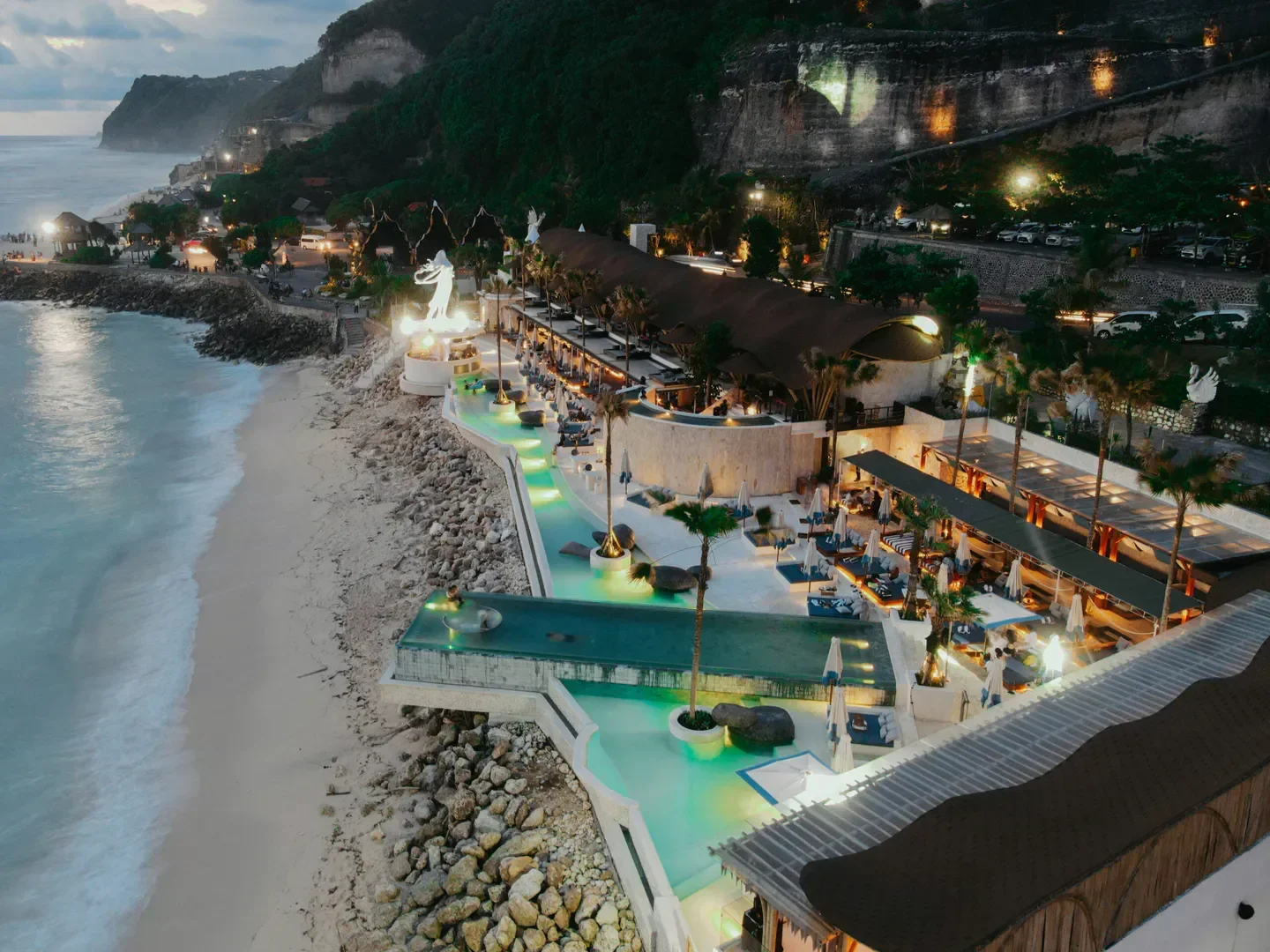 Nighttime aerial view by drone of a beachside resort featuring multiple swimming pools with turquoise lighting, lounge chairs with umbrellas, palm trees, a sandy shoreline, and cliffs in the background illuminated by various lights.