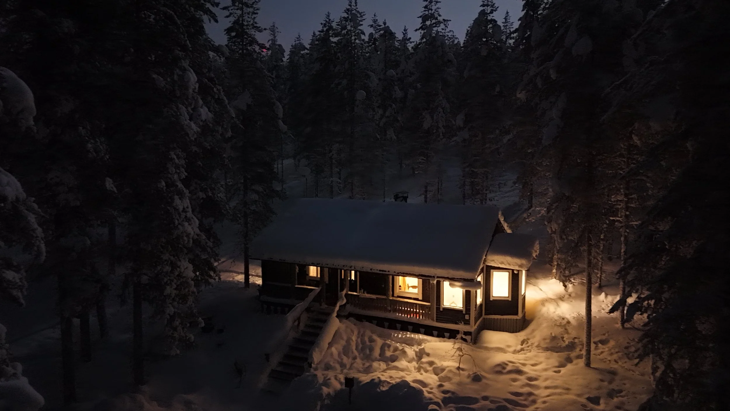 A snow-covered cabin at night in a forest, with warm light coming from the windows and a snow-covered staircase leading to the porch. Aerial view shot by drone.