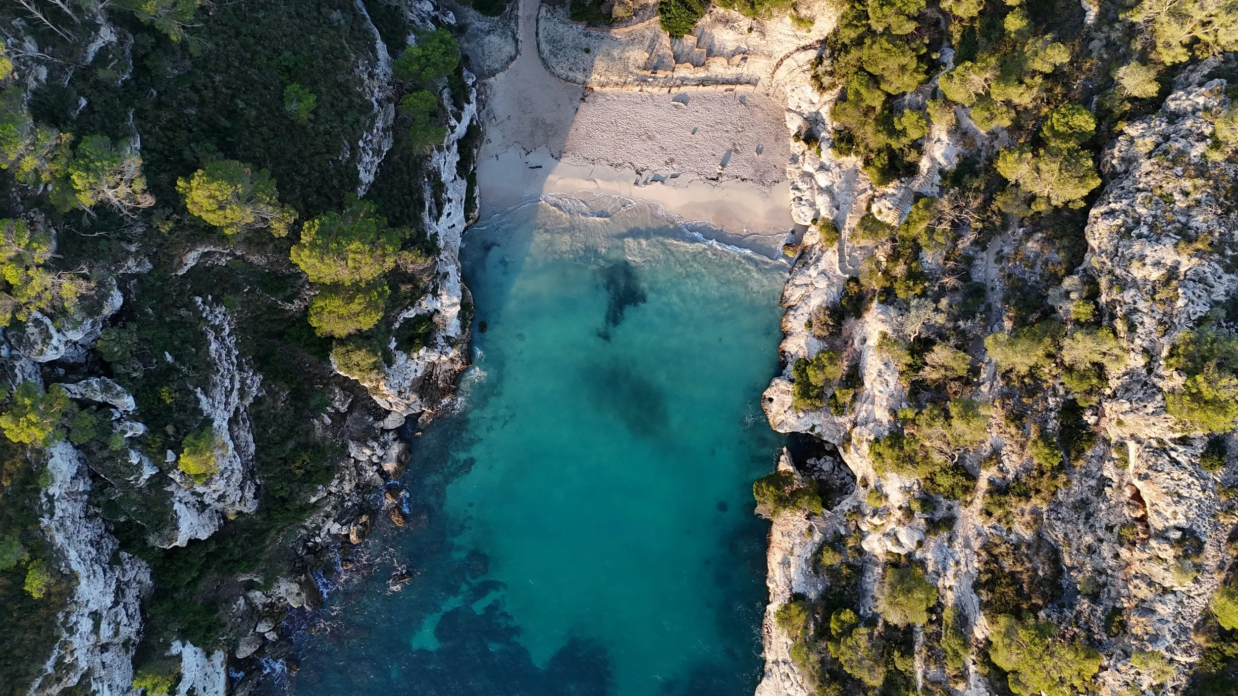 Aerial view shot by drone of a secluded cove with turquoise water, surrounded by rocky cliffs and green pine trees, featuring a small beach at the top of the cove. Cala Macarelleta in Menorca.