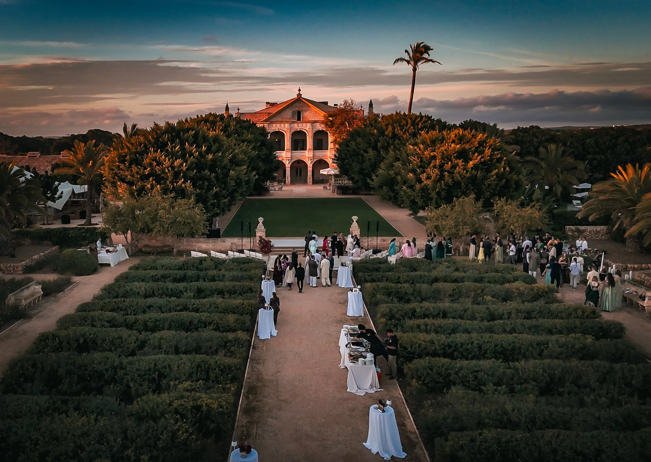 Drone aerial photography of an outdoor event at a large estate in Menorca with a grand mansion at sunset, guests mingling, and tables with white linens along a pathway.