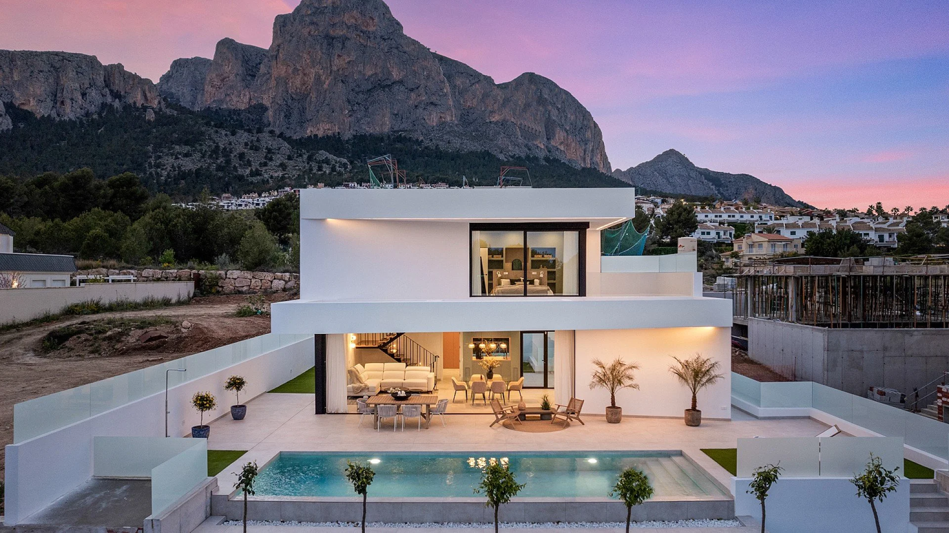 Drone aerial photography of A modern two-story white house in Menorca with large glass windows and doors, a swimming pool in the backyard, and outdoor seating, set against a mountain backdrop at sunset.