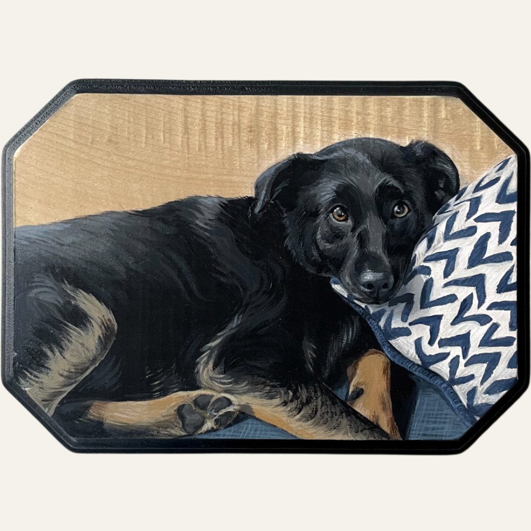 A black dog lying on a blue blanket, resting its head on a patterned pillow, with a wooden headboard in the background.