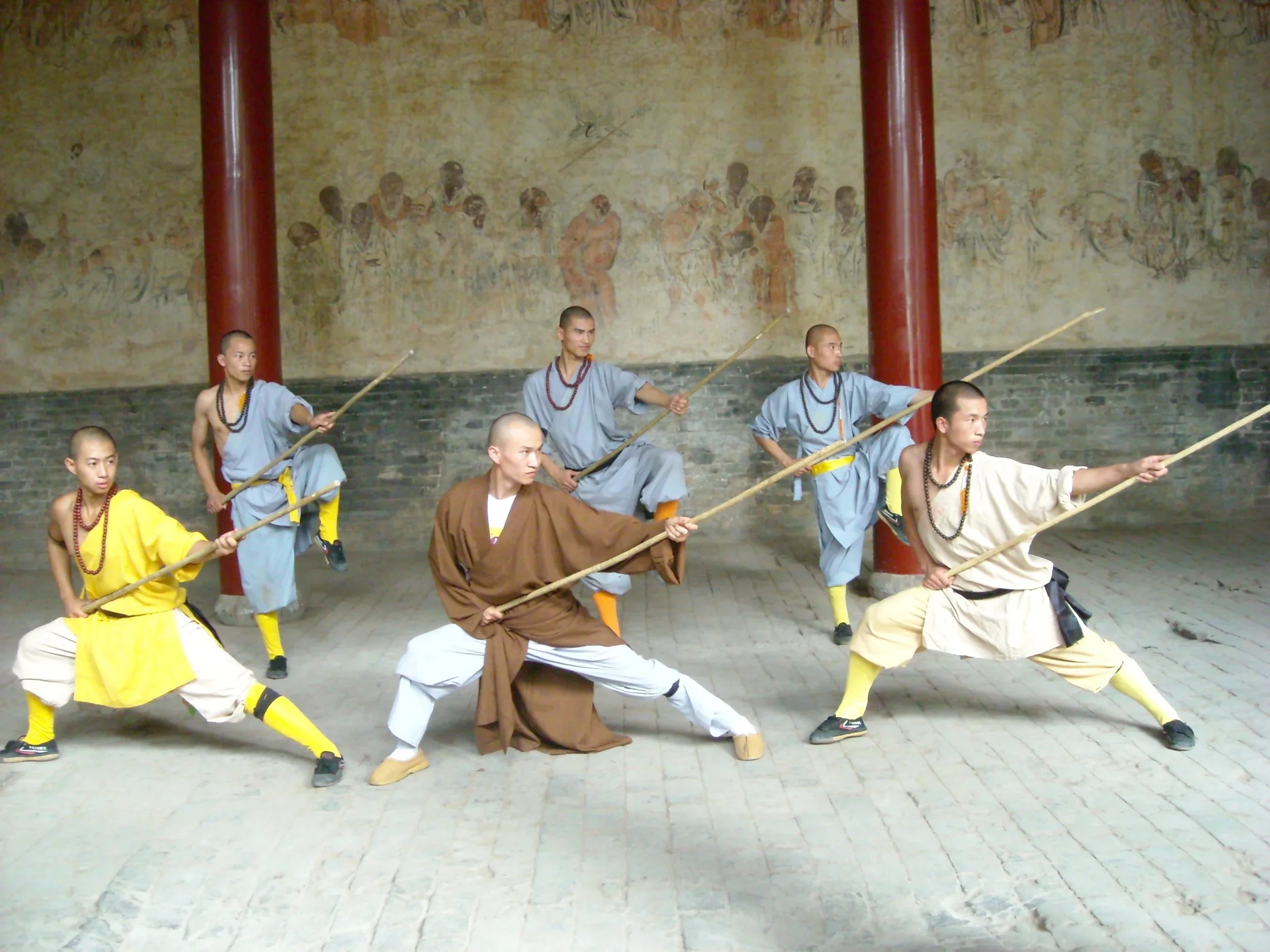 A group of martial artists practicing with staffs indoors, wearing traditional robes and yellow socks, with a mural on the wall behind them.