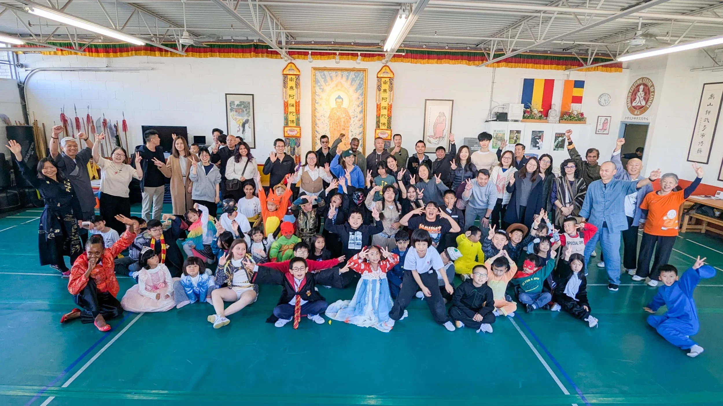 A large group of children and adults gathered in a gymnasium, some dressed in traditional Asian attire and others in casual clothing, celebrating together with many smiling and making peace signs.