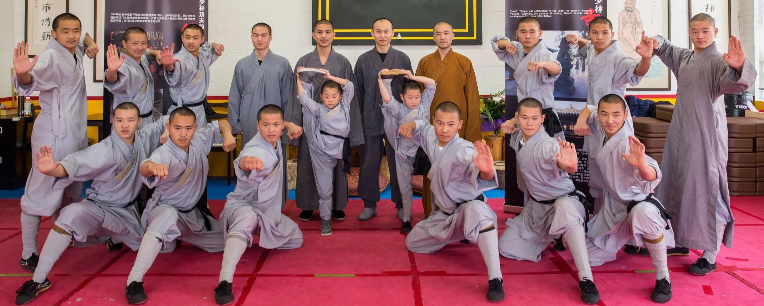 Group of martial arts students and instructors in uniform, performing martial arts poses inside a dojo or training room, with martial arts artwork on the walls.