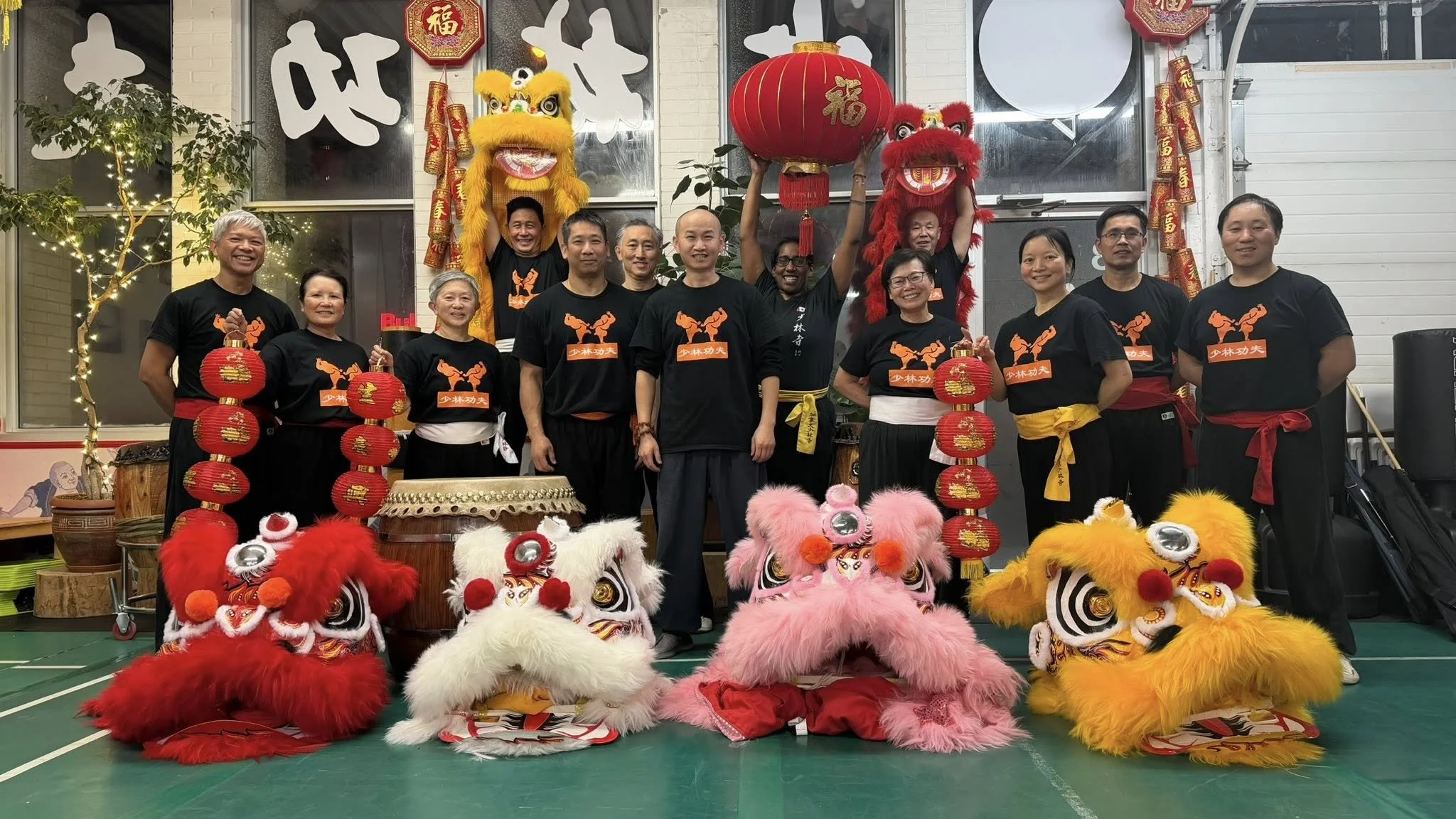 Group of people celebrating Chinese New Year with lion dance costumes, red lanterns, and decorative banners, indoors with festive decorations.