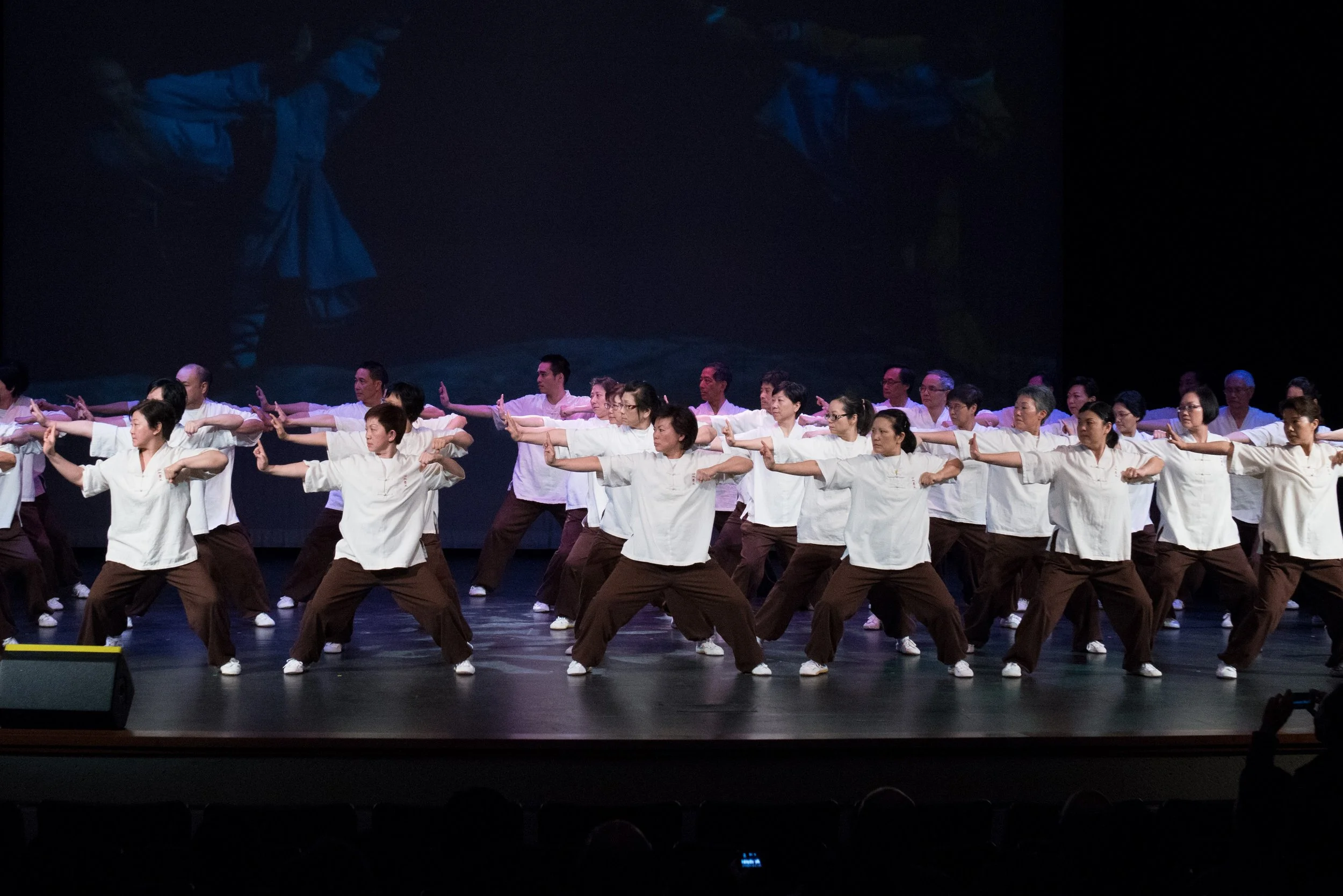 A group of people dressed in white shirts and brown pants performing a choreographed dance or Tai Chi on a stage with a dark background.
