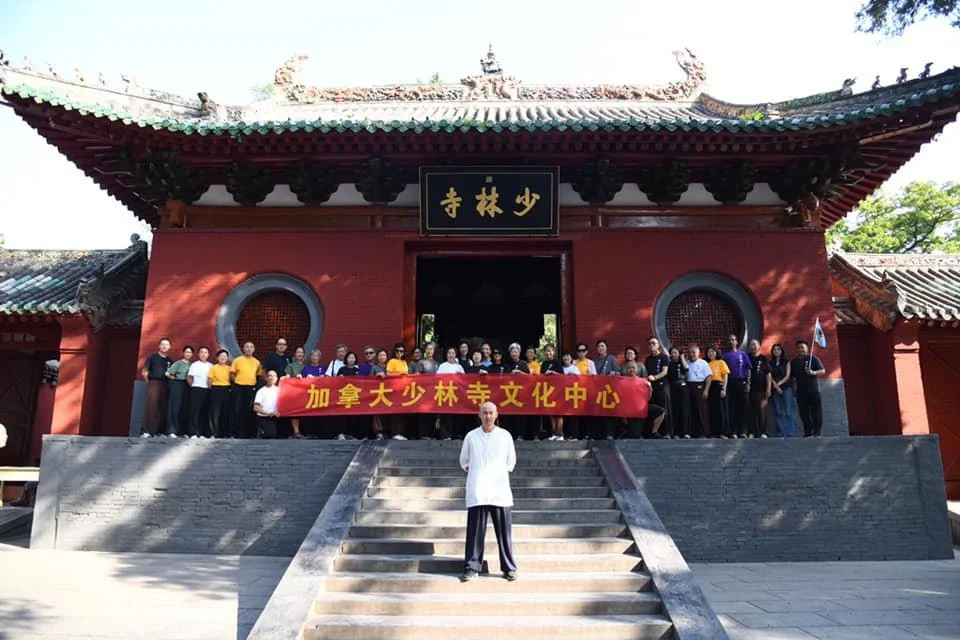 A large group of people standing on the steps of a traditional Chinese temple, holding a red banner with Chinese characters, with a man in traditional attire in front.