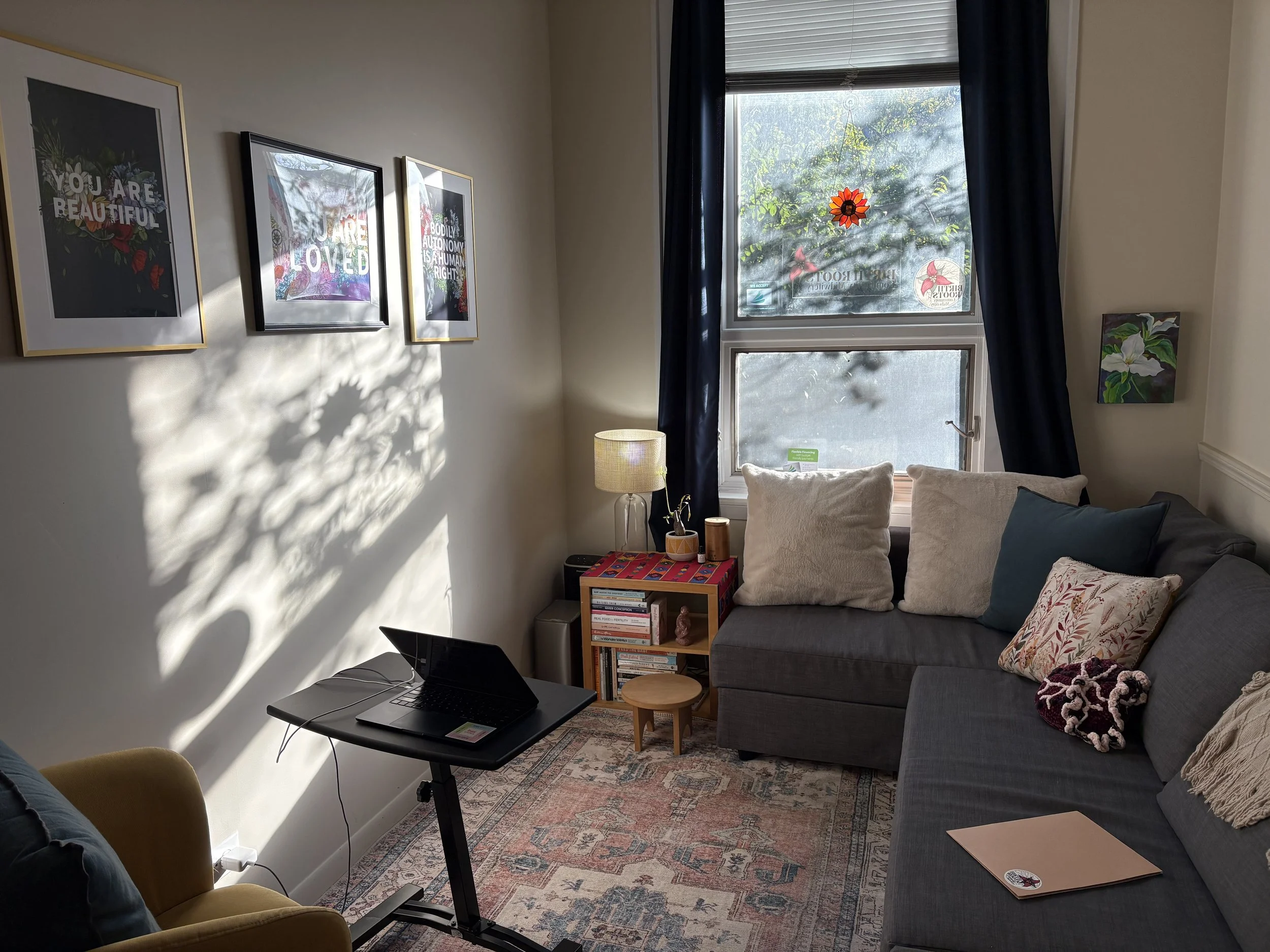 A cozy living room with a large window covered by dark curtains, sunlight casting shadows on the wall, and framed artwork. There is a gray sofa with decorative pillows, a small wooden side table, a patterned rug, a lamp, and a table with a laptop and other items.