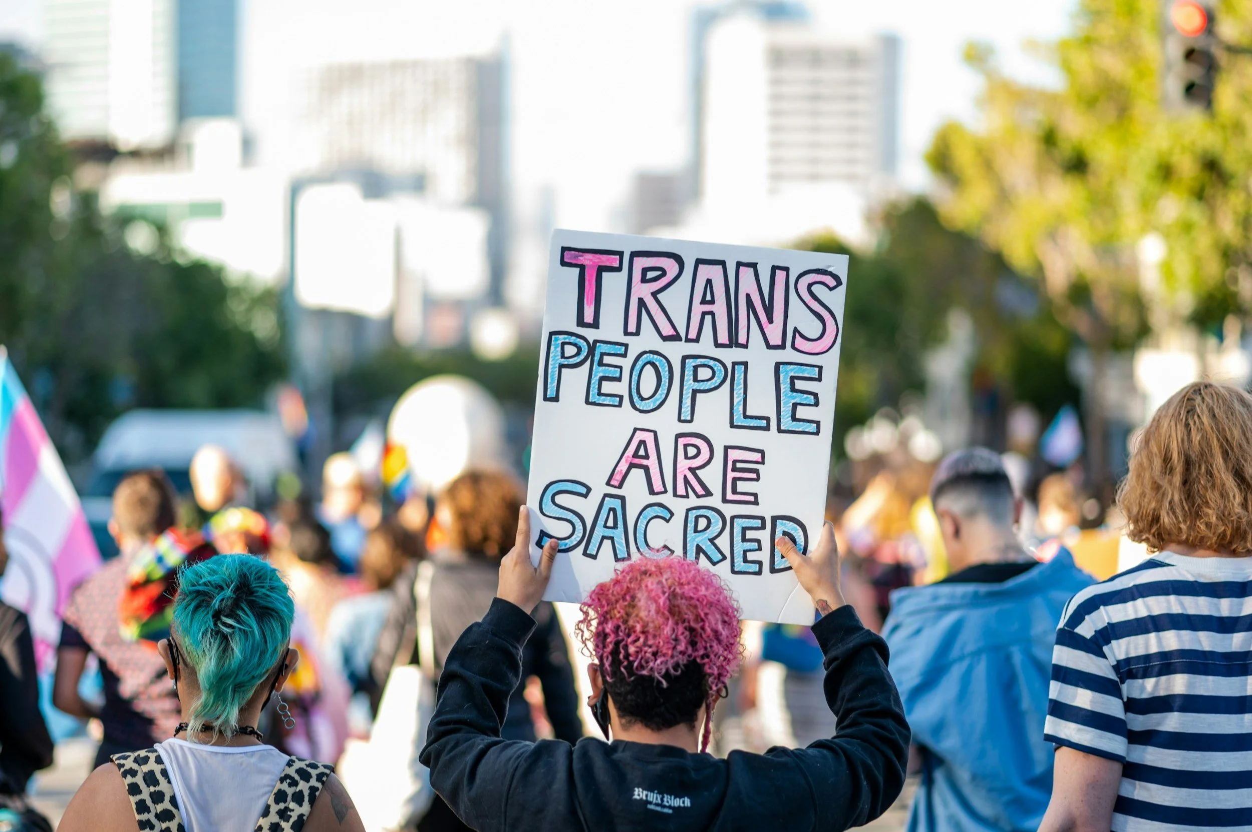 A crowd of people participating in a pride parade, with one person holding a sign that reads 'Trans People Are Sacred' in colorful letters. The crowd includes individuals with vibrant hair colors and casual clothing, and the background features city buildings and trees.