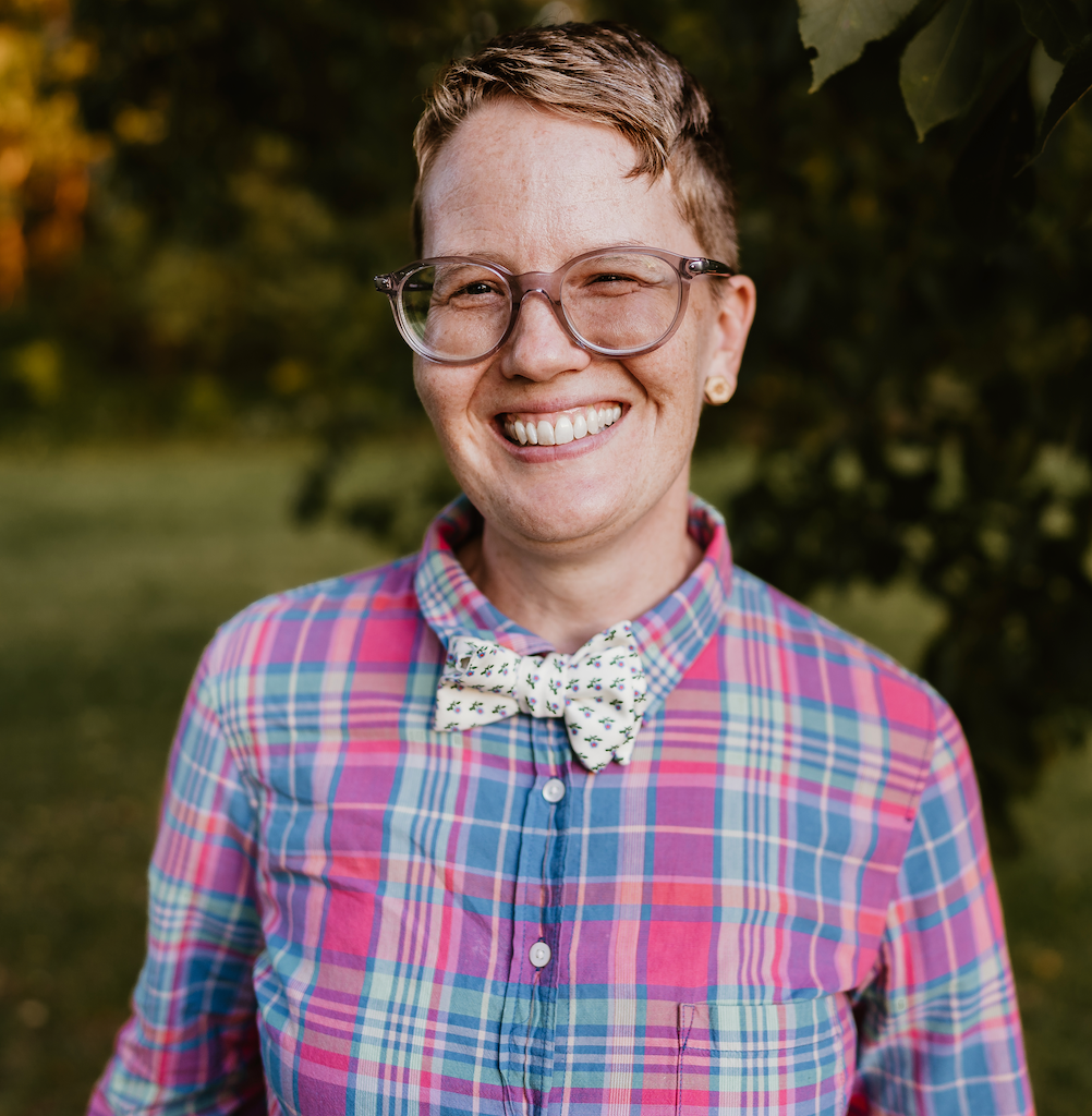 smiling Corinne in short, masc haircut, wearing a pastel plaid shirt and white flowered bowtie