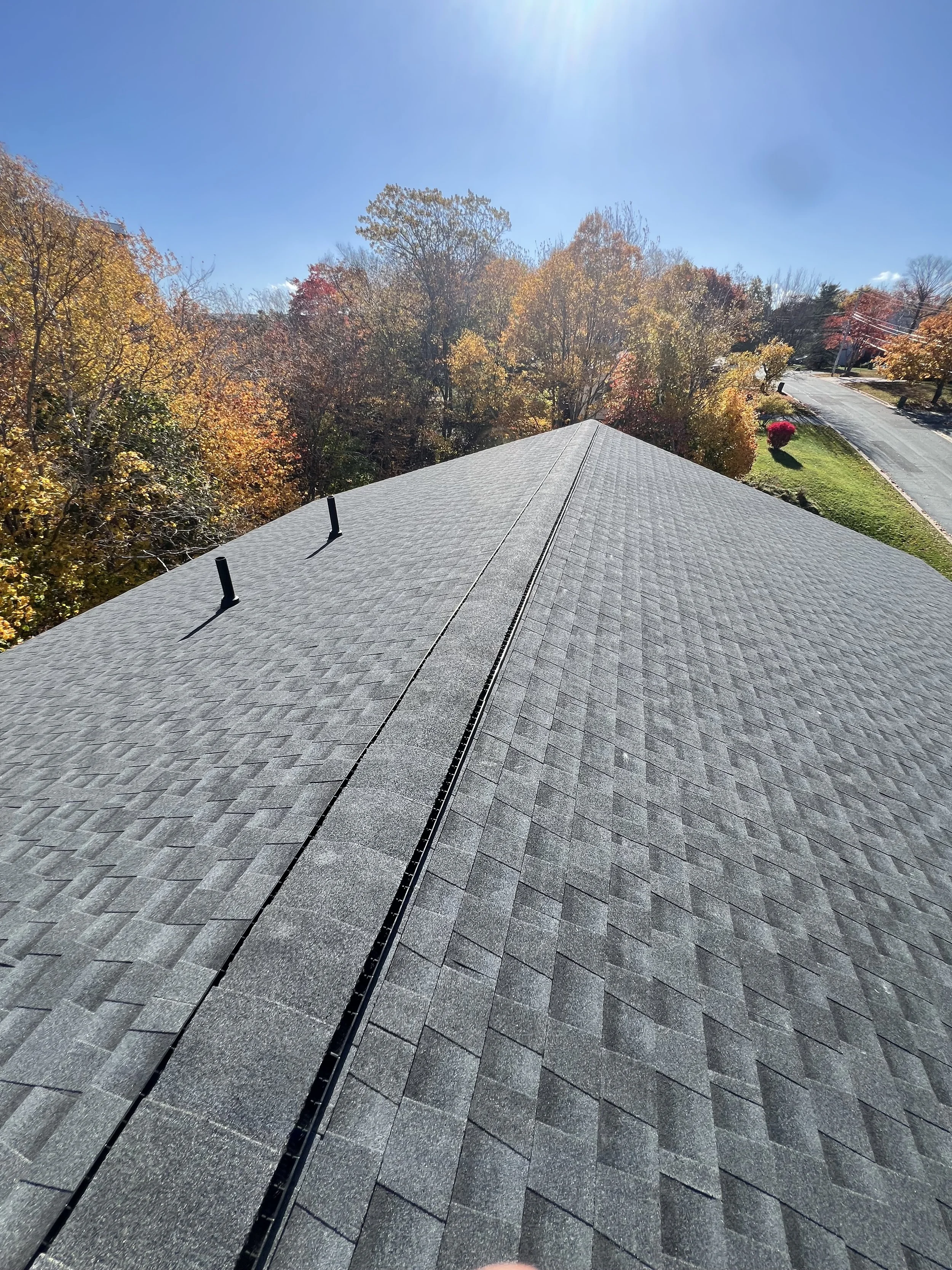View of the roof of a house with shingles, showing two vent pipes and a middle ridge vent. In the background, there are colorful fall trees, a sidewalk, and a street with a lawn and a red bush.