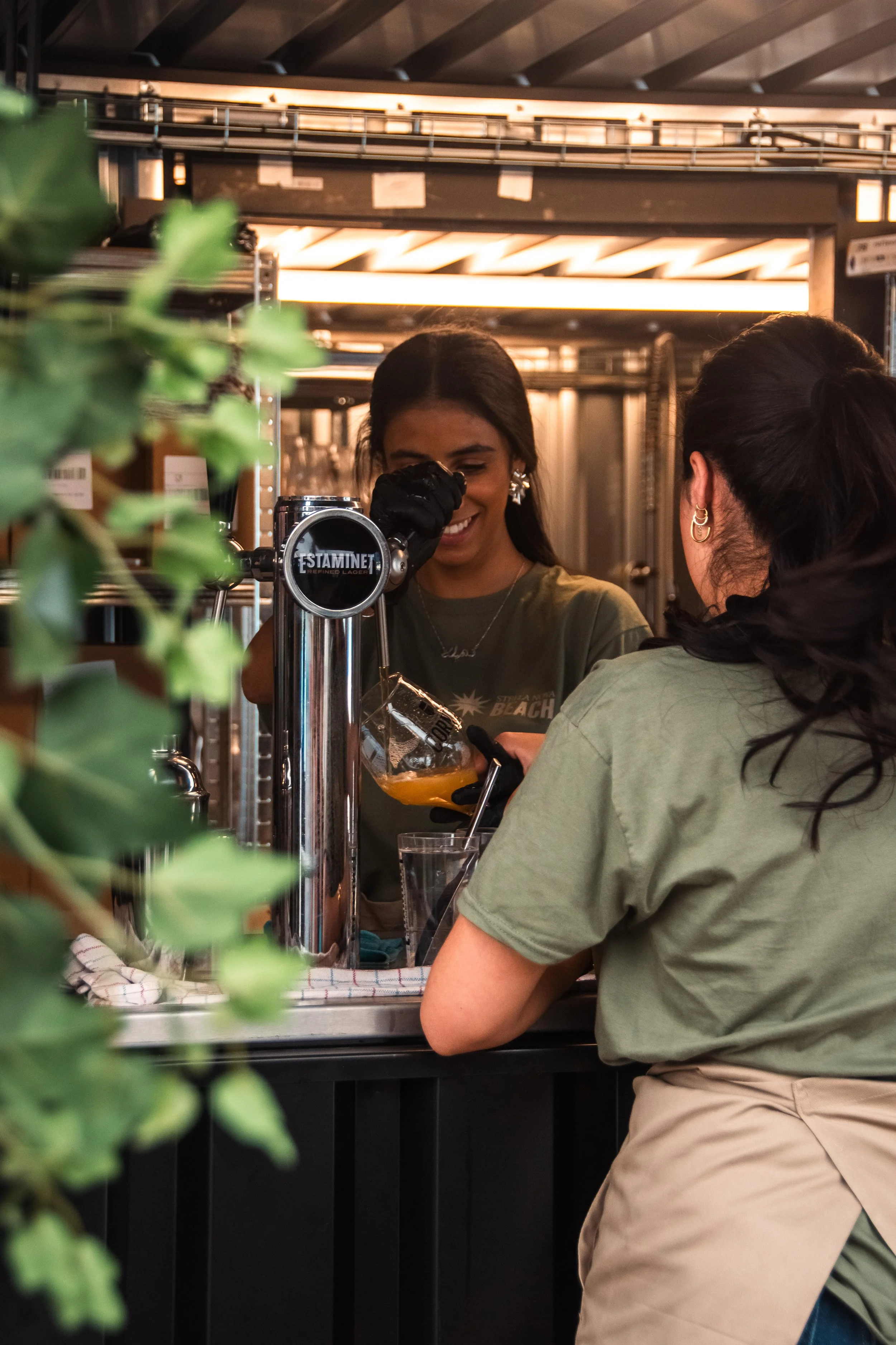 Two women are serving drinks at a bar, one woman is holding a pitcher and pouring a beverage into a glass, while the other woman is facing away. The bar area has metal shelves and warm lighting, with some green foliage in the foreground.