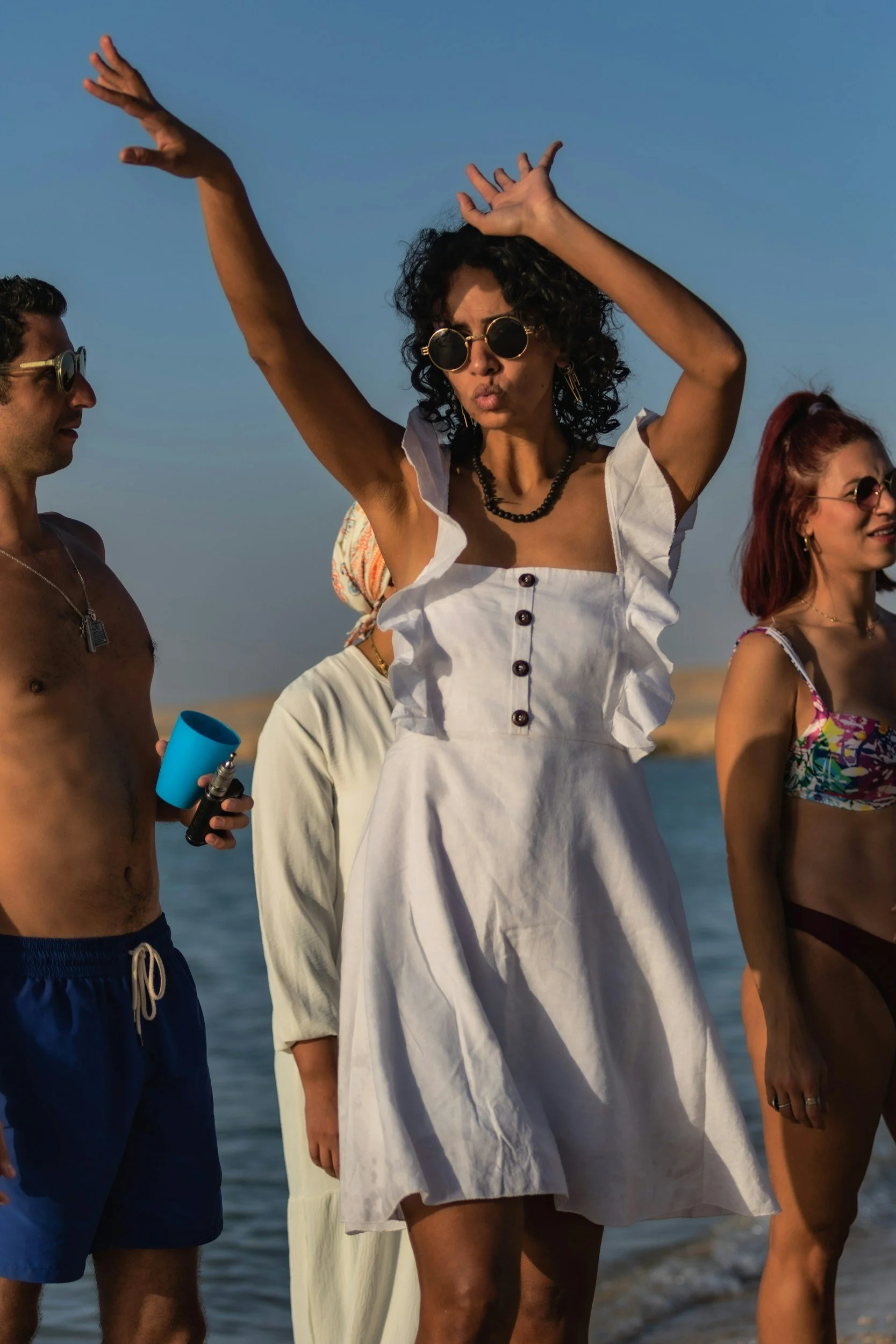 A group of people at the beach, with a woman in the center wearing sunglasses and a white dress, raising her arms, and others around her wearing swimwear and sunglasses.