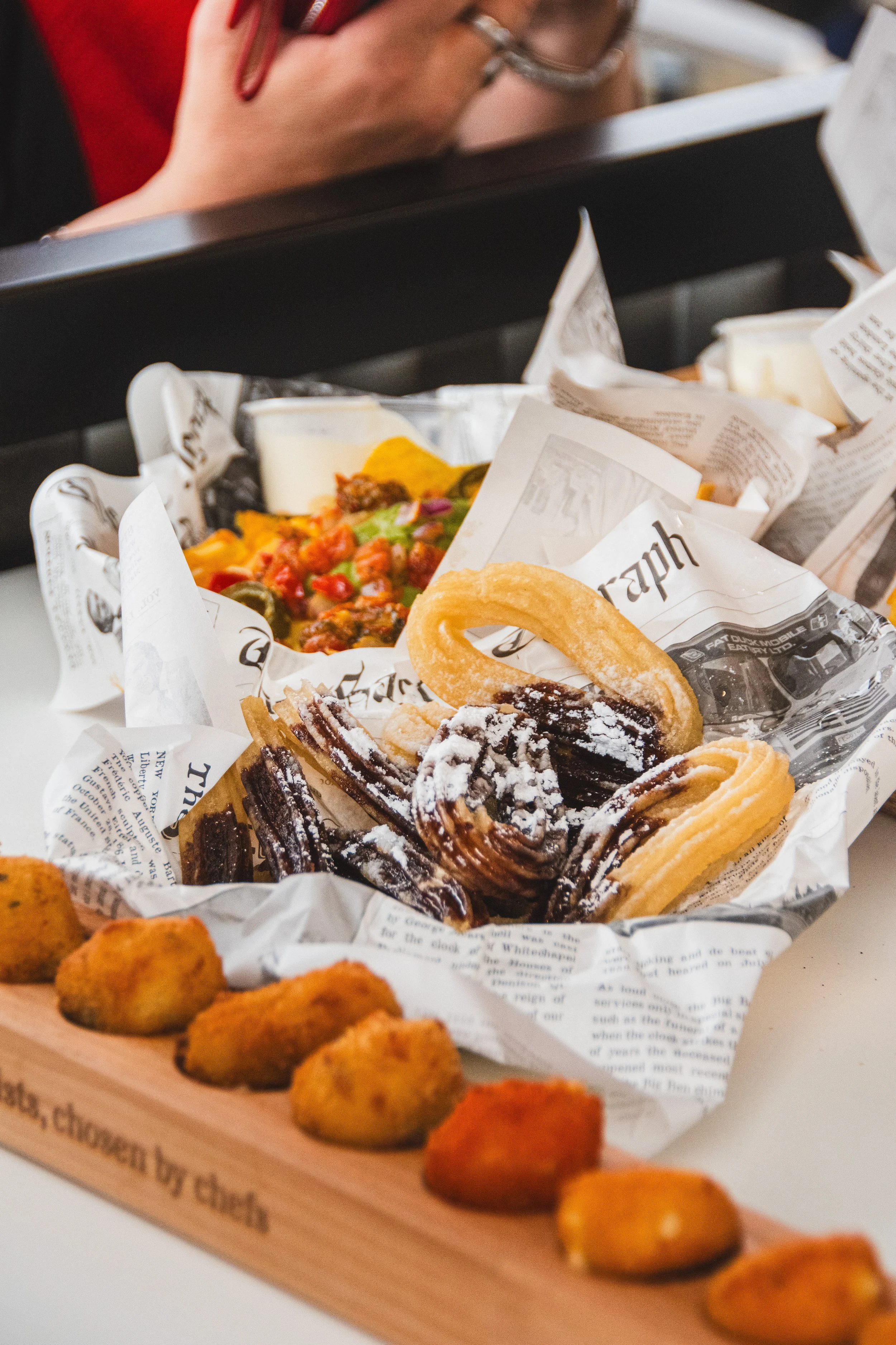 A tray of assorted fried foods, including mozzarella sticks, onion rings, and fried chicken nuggets, with a black basket containing a salad, dressing, and other food items in the background.
