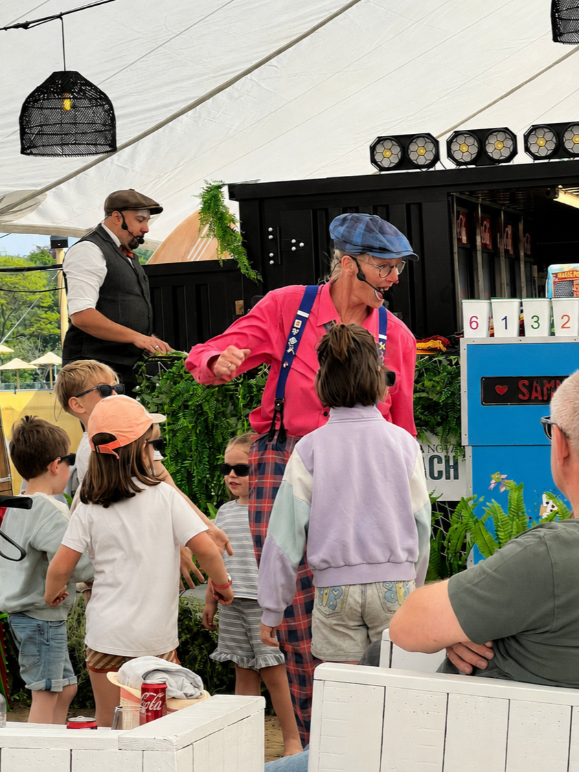 A children's magic show under a large tent with a performer in colorful attire and an audience of kids wearing sunglasses.